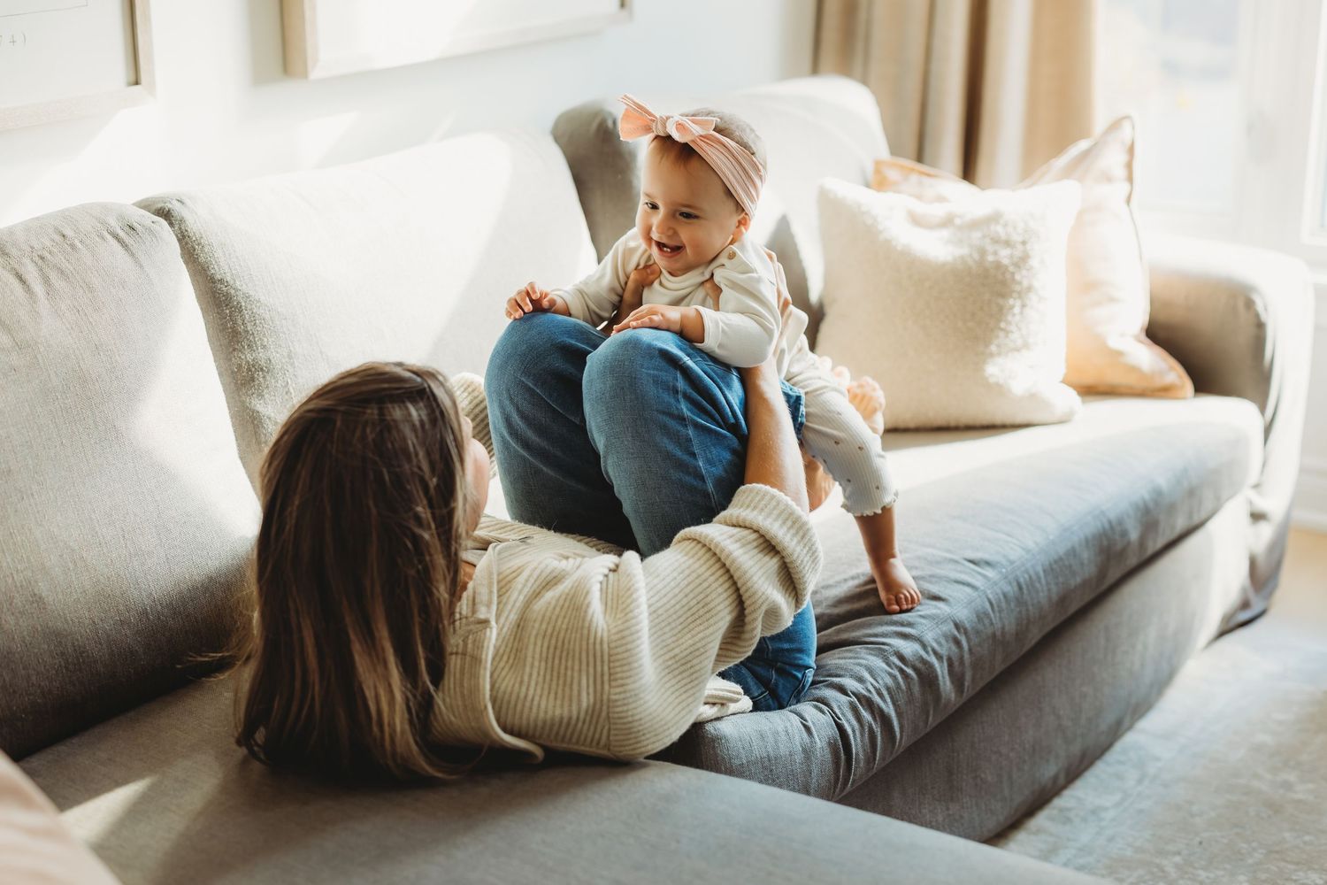 A mother playfully holds her baby on her knees while sitting on a cozy gray sofa in a bright living room.