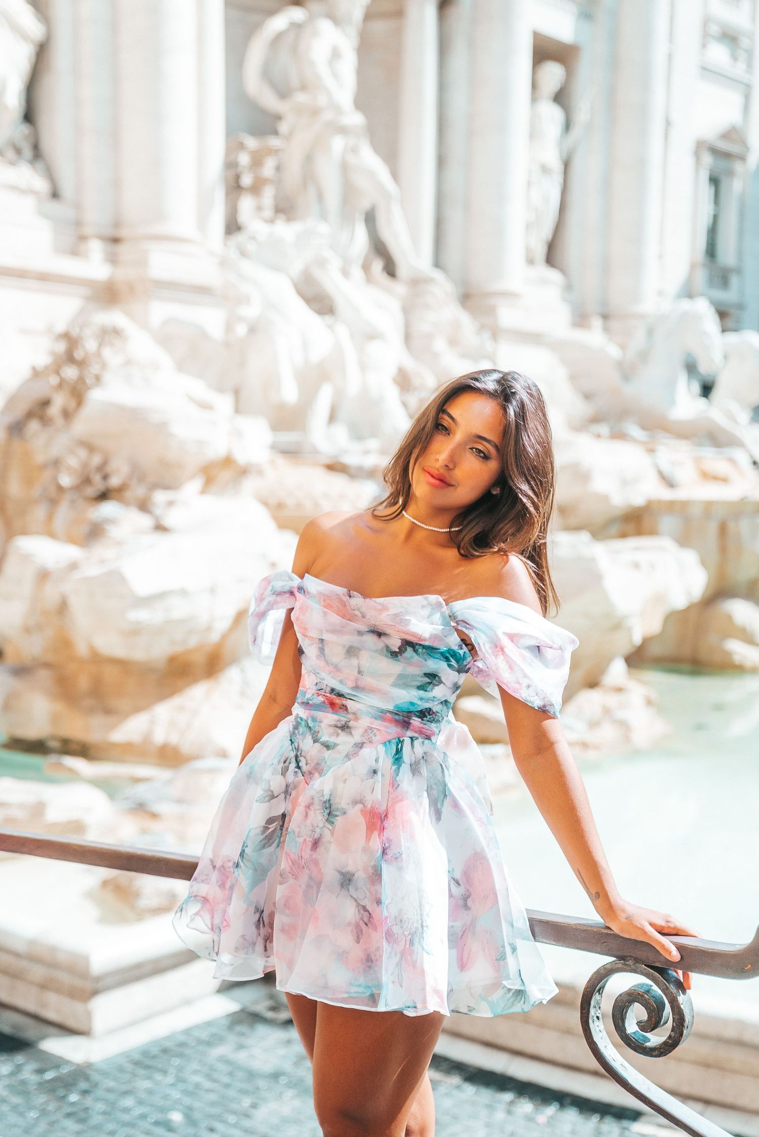 A person in a flowy floral dress poses playfully near a historic fountain in a romantic travel photo series.