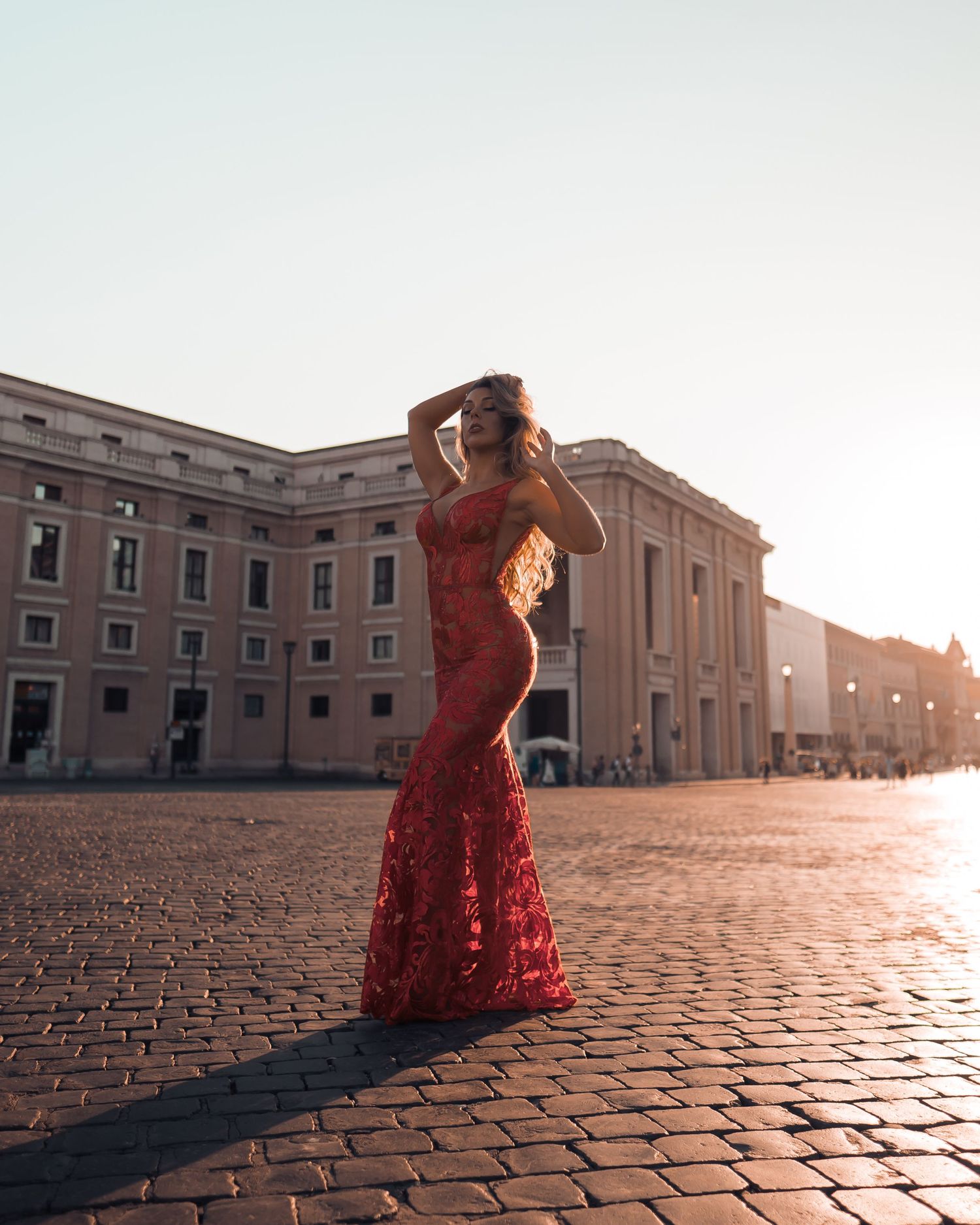A figure in a flowing red dress poses gracefully on cobblestone pavement at sunset near historic architecture.