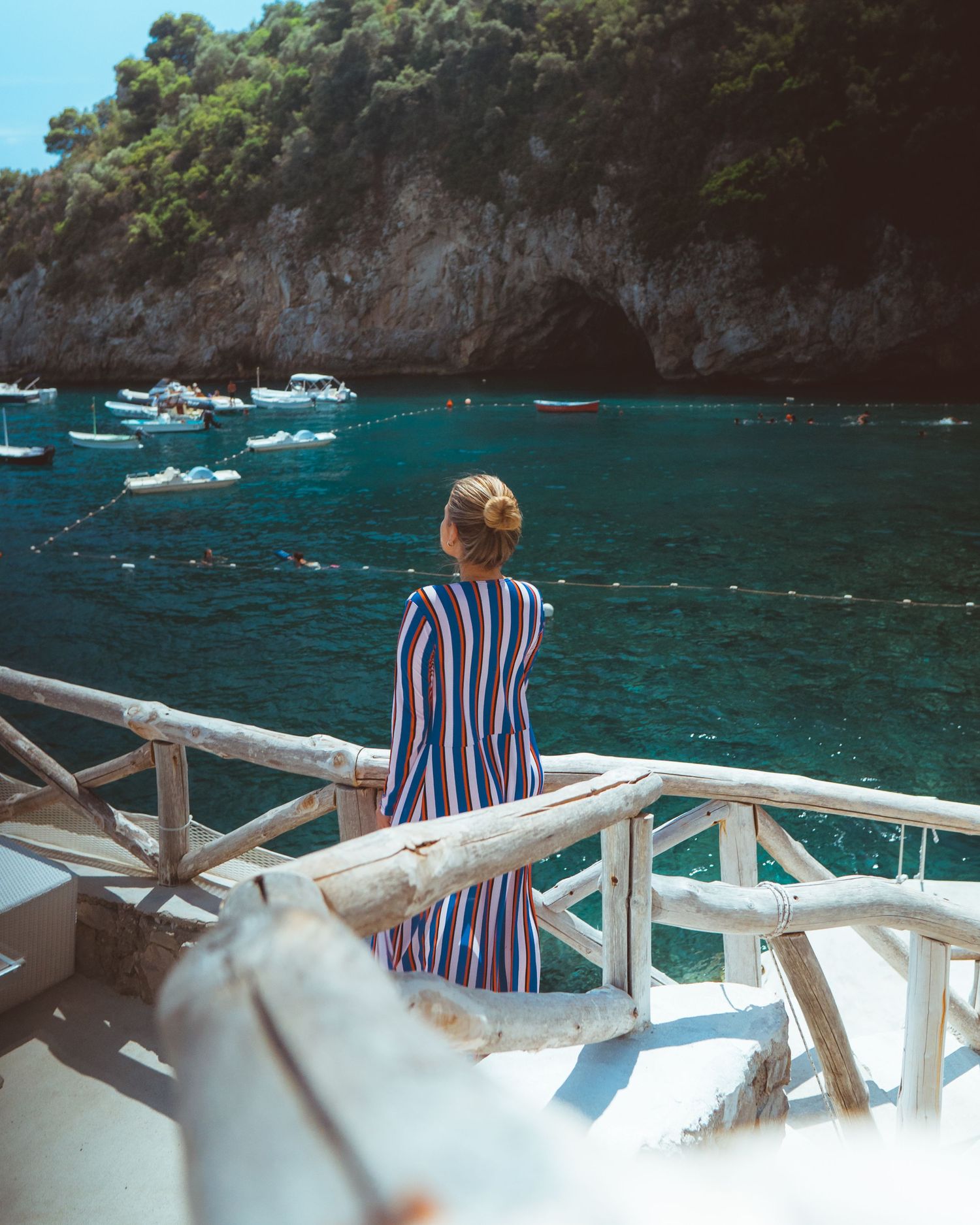 Person in striped shirt sits on white boat railing overlooking crystal blue Mediterranean waters and rocky coastline.