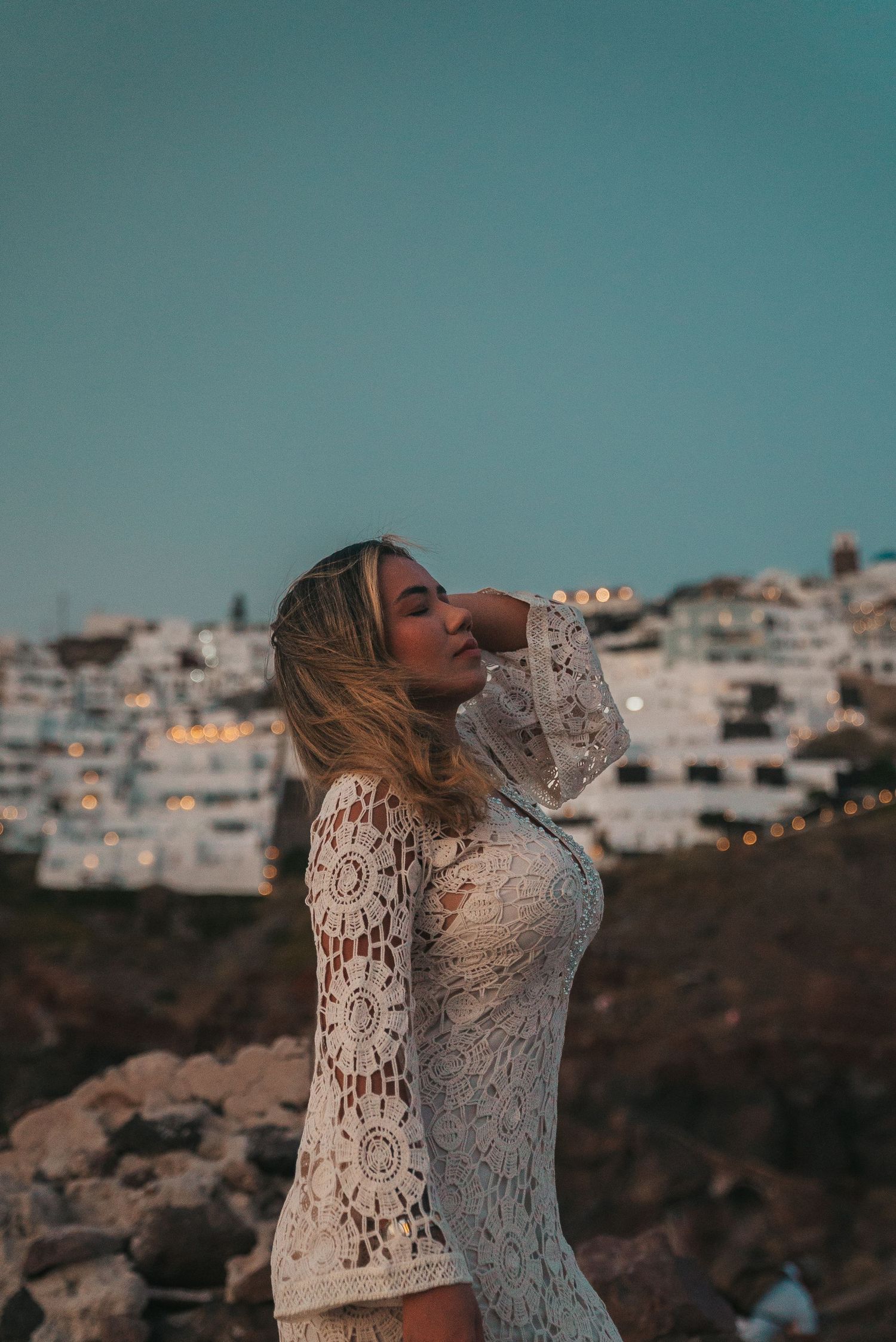 Person in white lace dress poses against coastal cityscape backdrop during golden hour.
