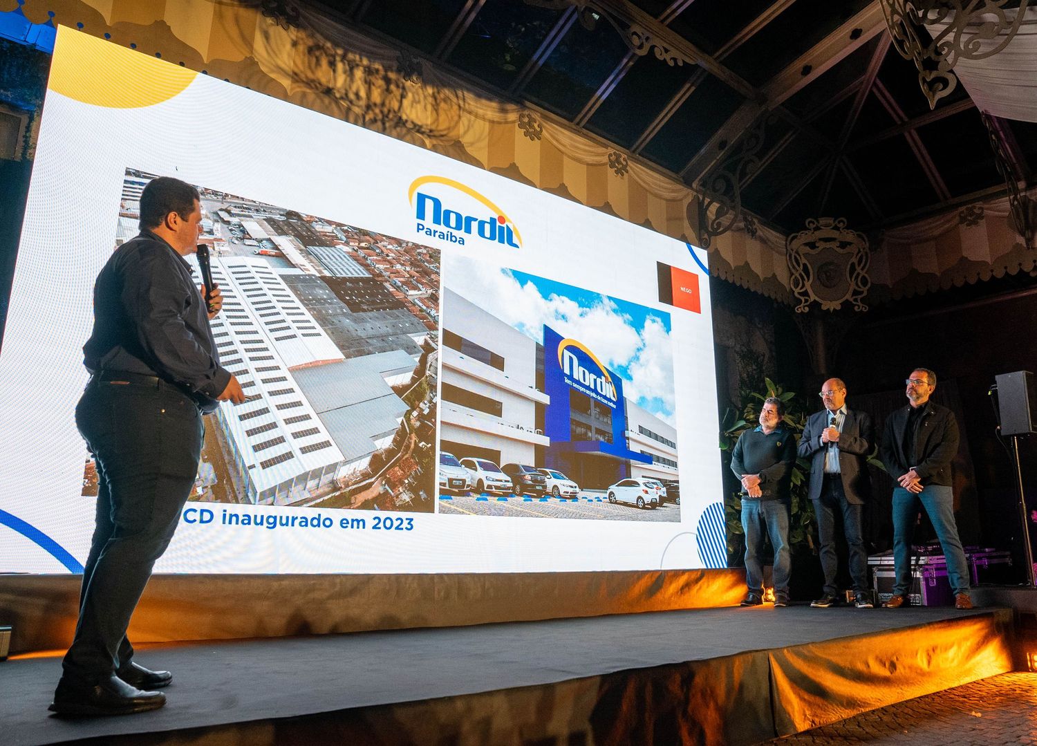 A panoramic view of a presentation stage with large projection screens displaying business content in a dark room.