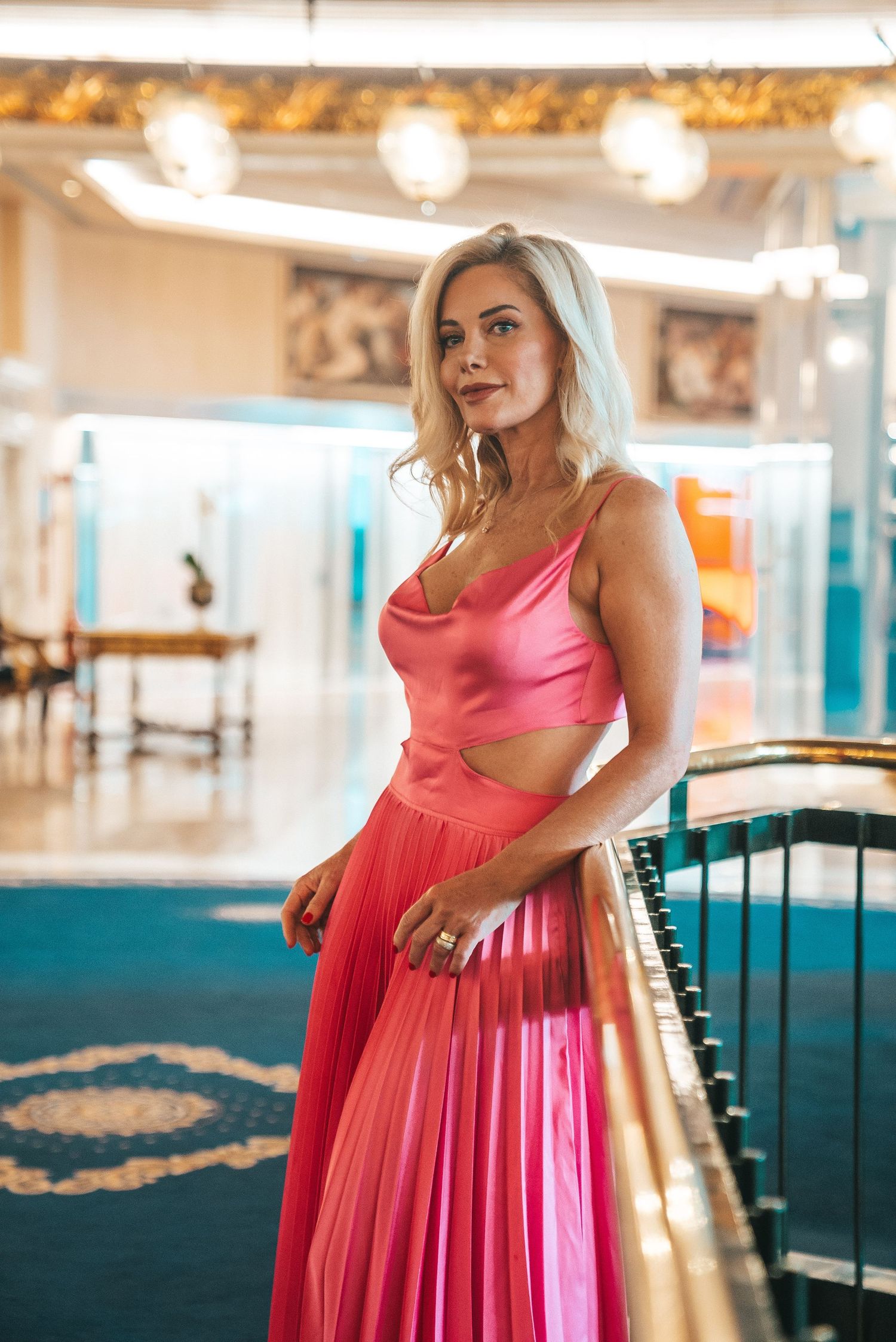 A woman in a coral pink dress poses elegantly in an ornately decorated room with vintage decor.