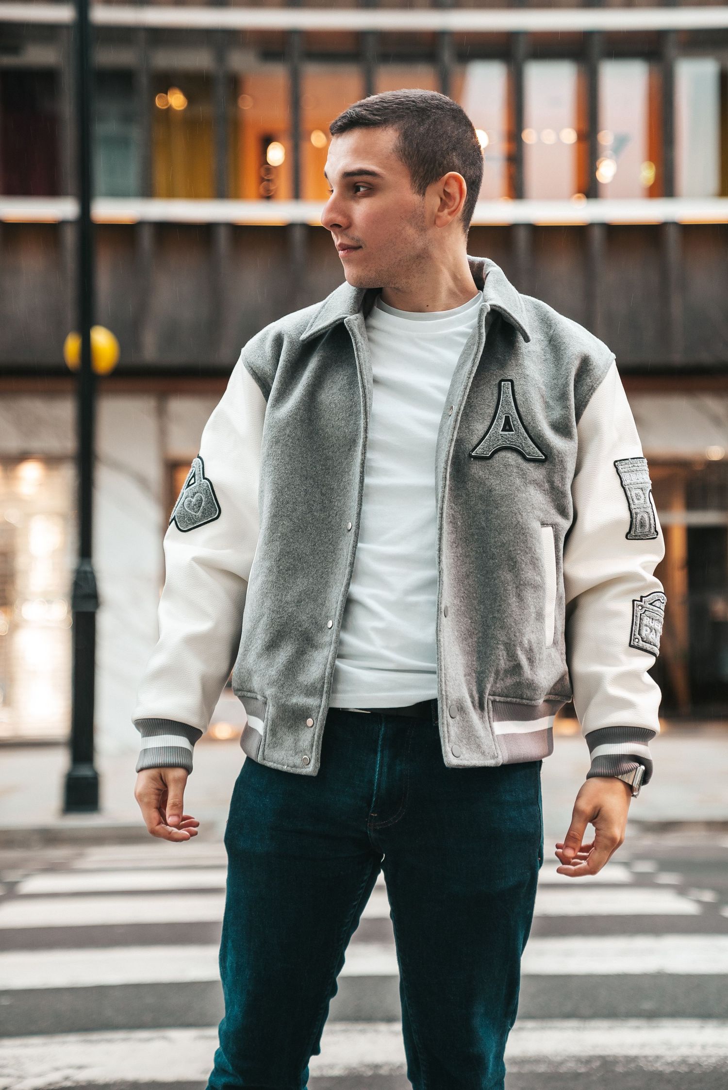A person wearing a grey and white varsity jacket with dark jeans poses on a city street.