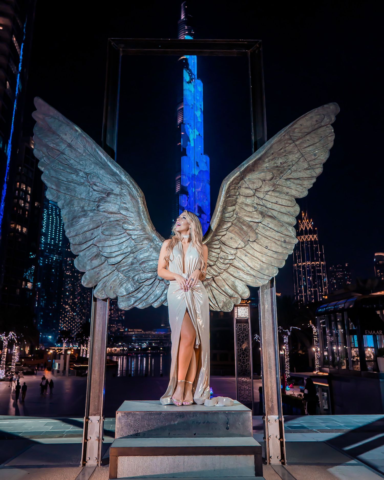 A figure in a white dress stands on a platform with illuminated angel wings against a dark cityscape at night.