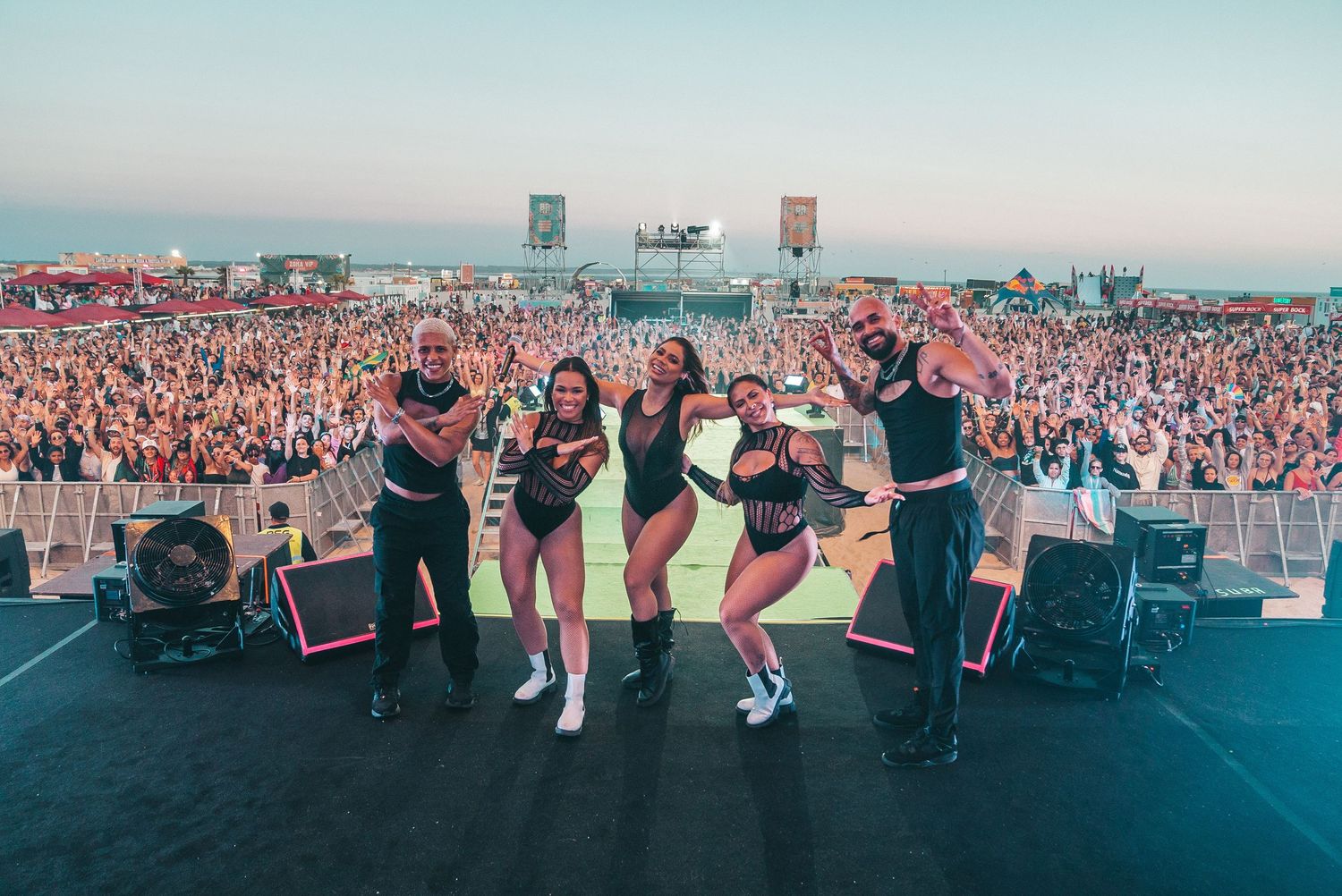 Performers on stage energetically entertain a massive crowd at an outdoor music festival during sunset.