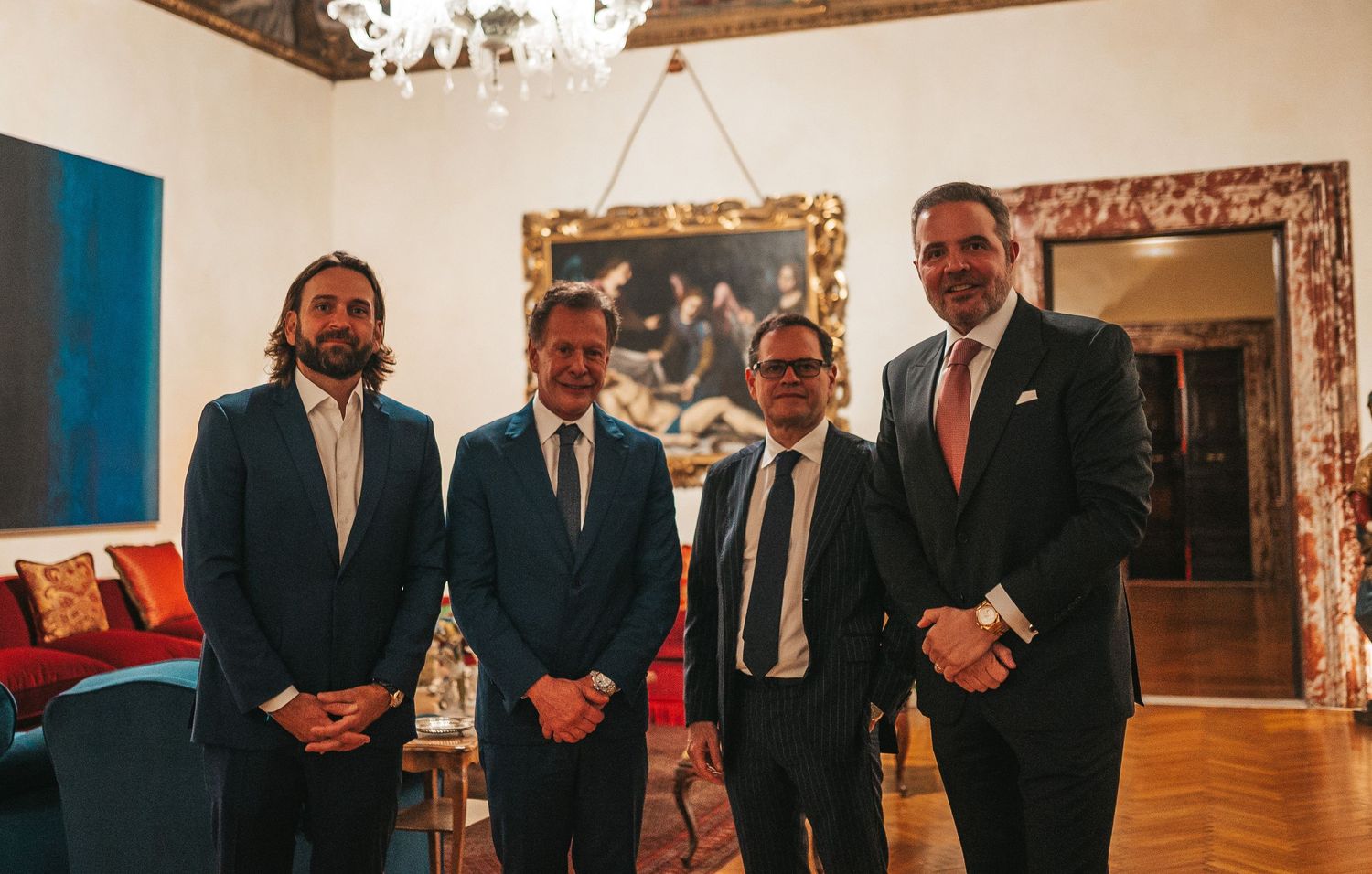 A group of formally dressed people stand in a row inside an elegant room with chandeliers and framed artwork.
