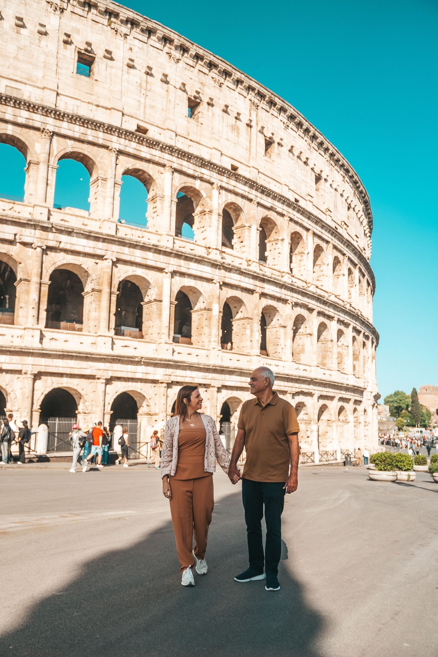 Two people walking along the exterior of the Colosseum on a sunny day with bright blue skies.