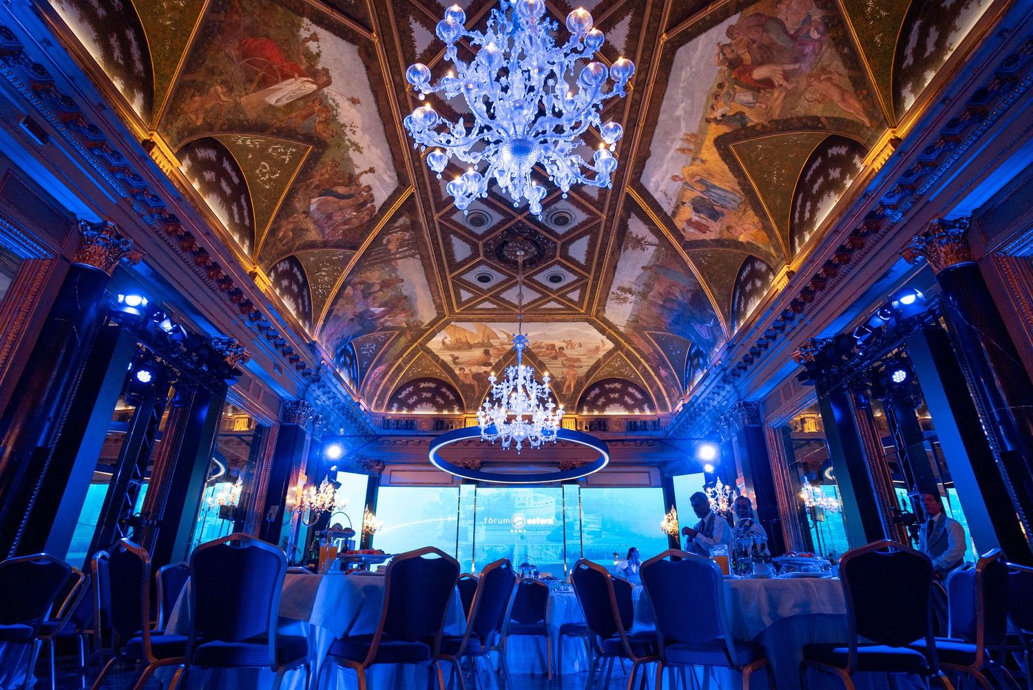 Elegant ballroom with ornate chandeliers and blue uplighting creates a dramatic atmosphere for special events.
