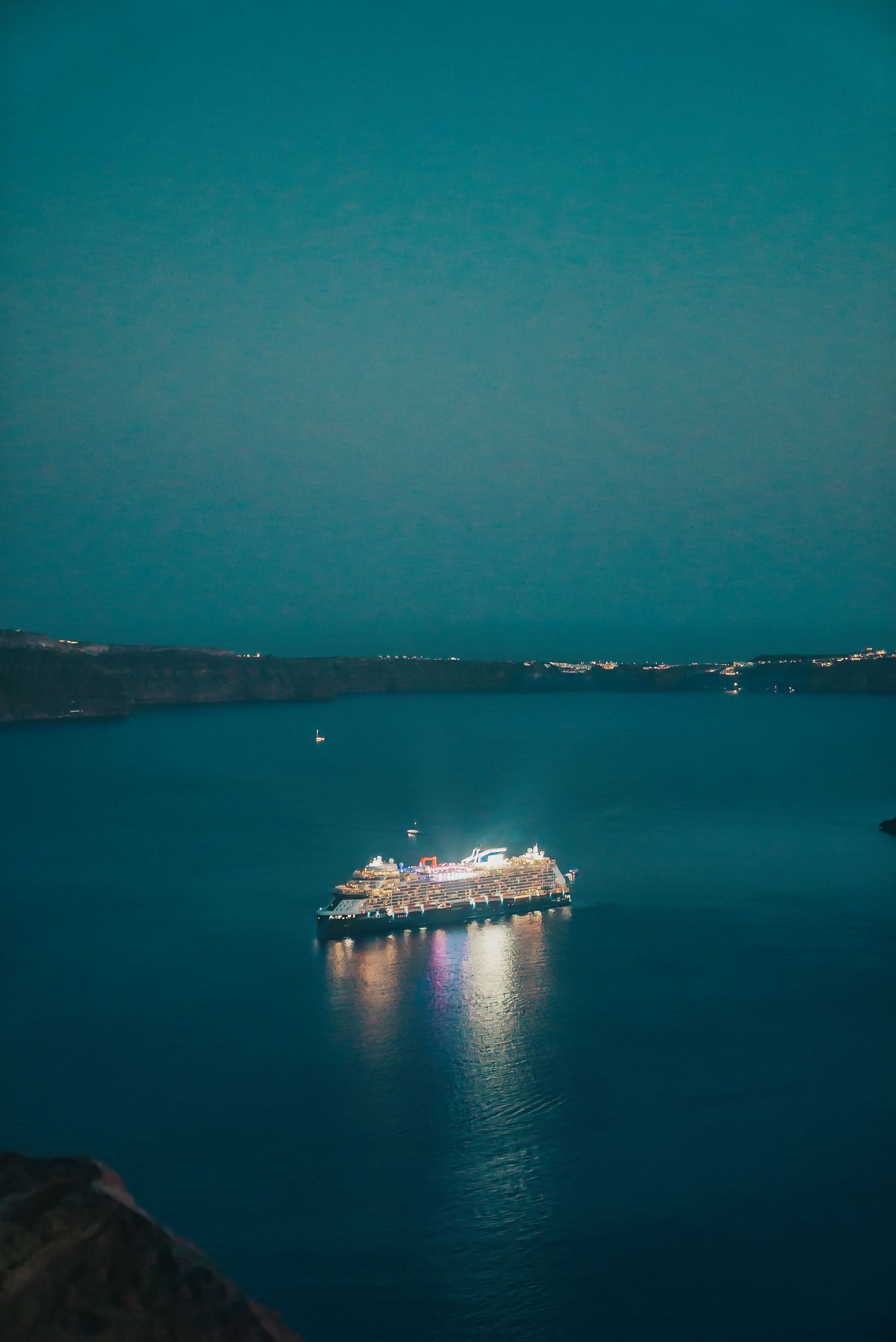 Illuminated cruise ship glows against dark turquoise waters at night with distant shoreline silhouetted in the background.