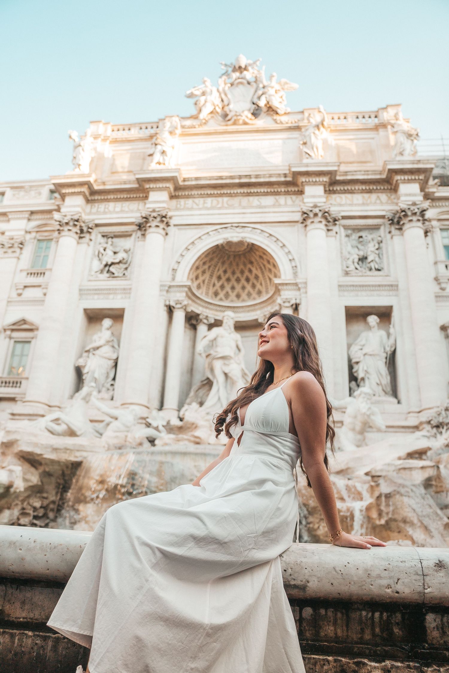 Elegant white dress flowing in front of the iconic Trevi Fountain in Rome during sunrise.