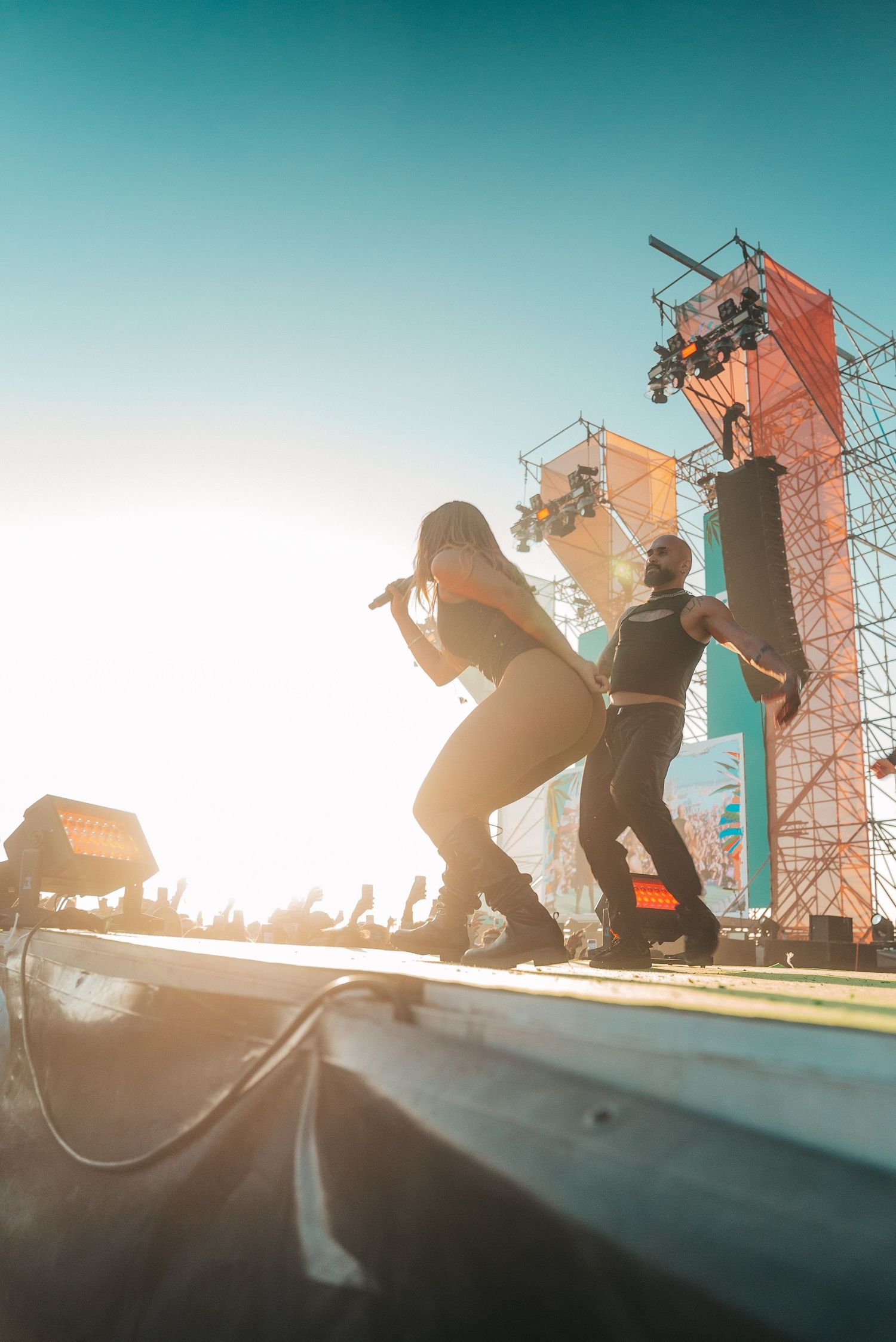 Silhouetted performers on stage at an outdoor music festival with large speaker towers against a turquoise sunset sky.