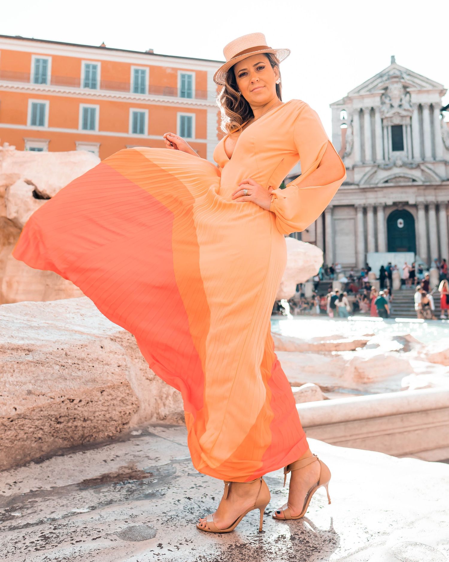 A woman in a flowing orange dress and sun hat poses gracefully in front of historic European architecture.