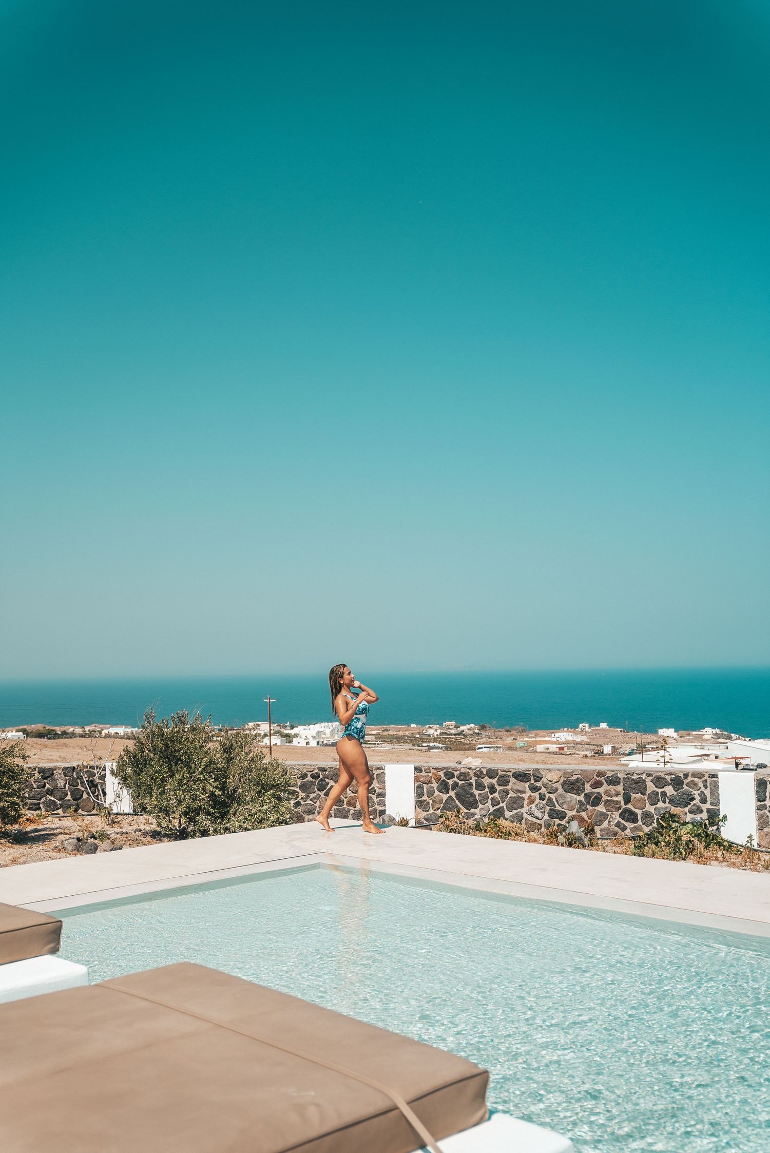 Infinity pool overlooking coastal landscape with turquoise water and white buildings against bright blue sky.