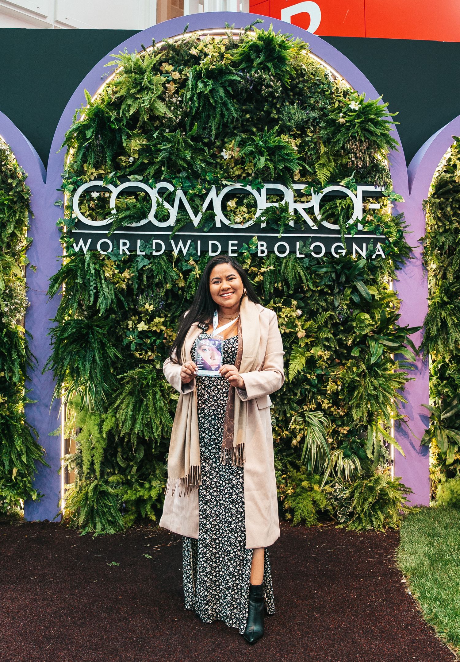 Person posing in front of a green leaf wall display with Cosmoprof Worldwide Bologna branding.