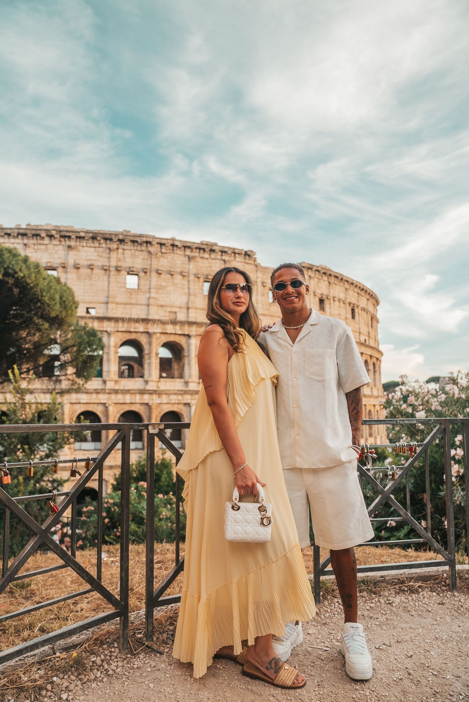 Couple posing in front of the Colosseum in Rome, woman in yellow dress, man in white outfit on a sunny day.