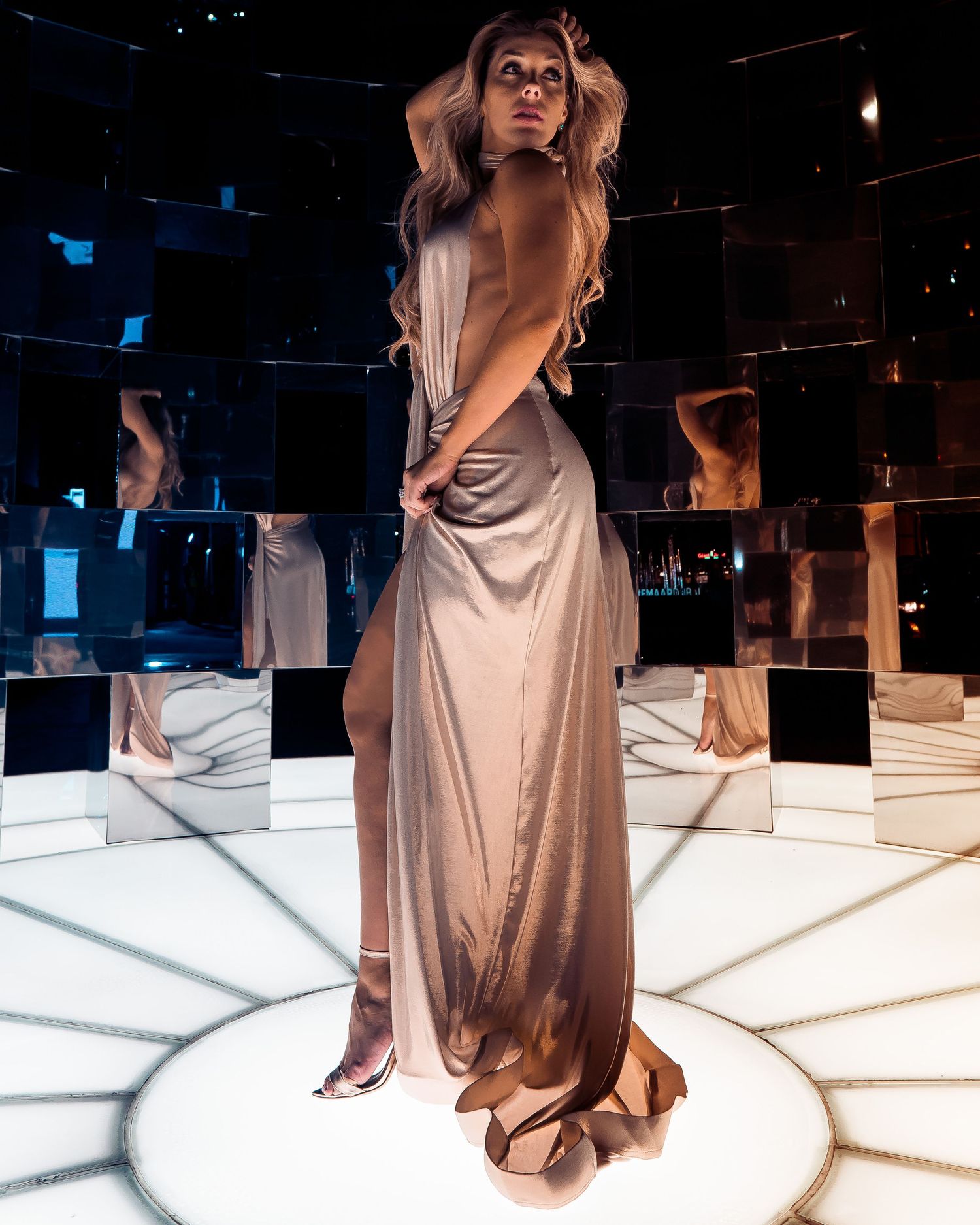 A model in a flowing beige dress poses dramatically against a dark background with illuminated floor panels.