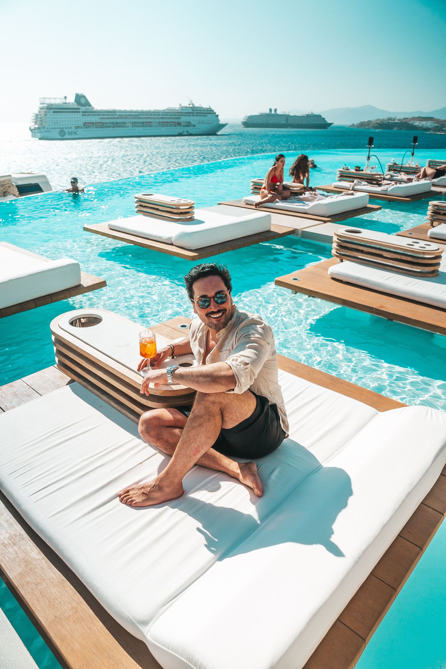 Luxury poolside lounge at a Mediterranean resort with cruise ship visible in background and turquoise infinity pool waters.