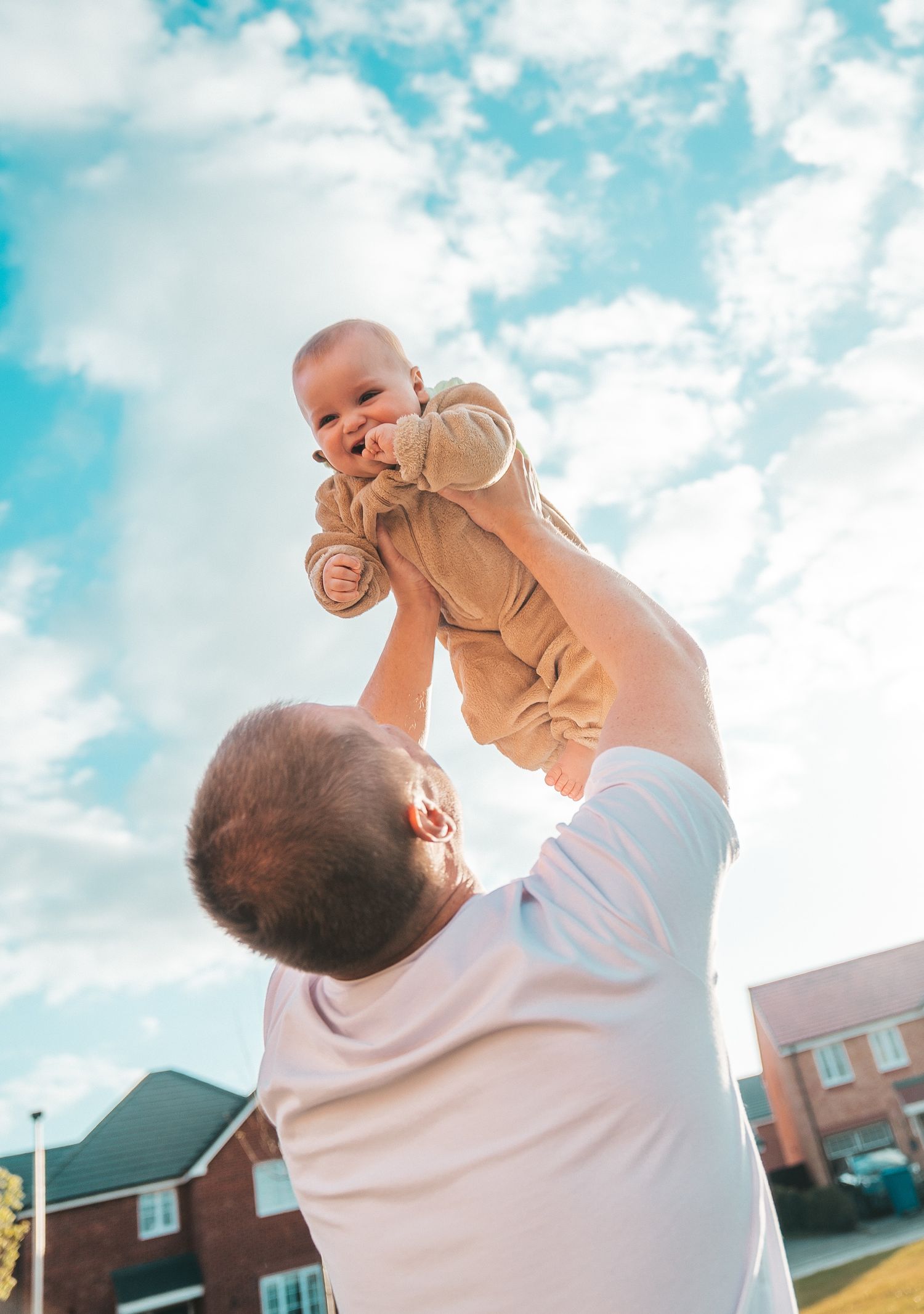 A joyful moment captured against a bright blue sky with fluffy clouds as a baby is lifted high into the air.