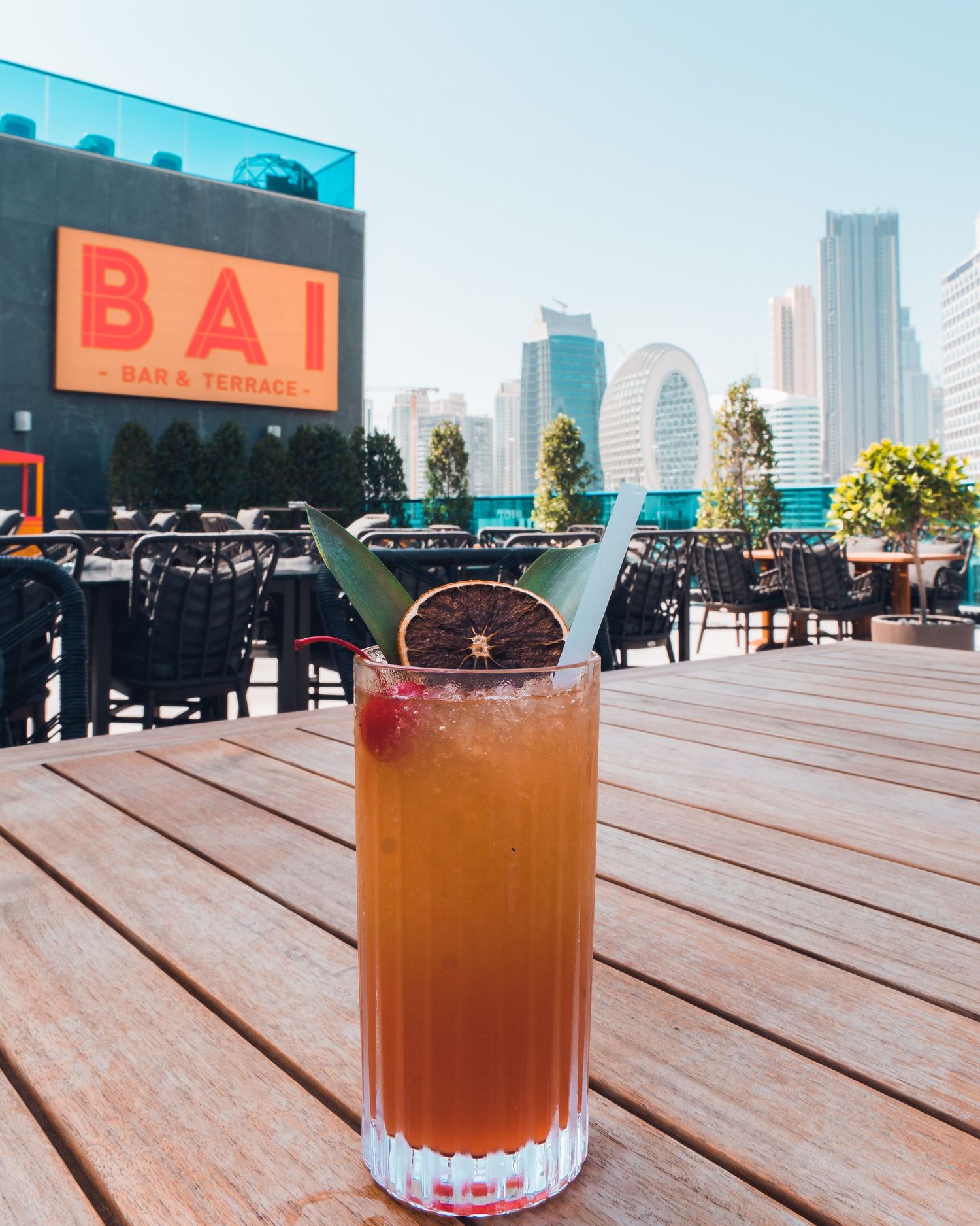 An orange cocktail in a crystal glass sits on a wooden deck with city buildings visible in the background.