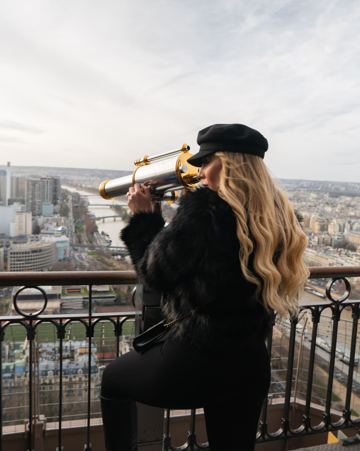 Person in black coat and hat looking through telescope at Paris cityscape from high viewpoint on cloudy day.