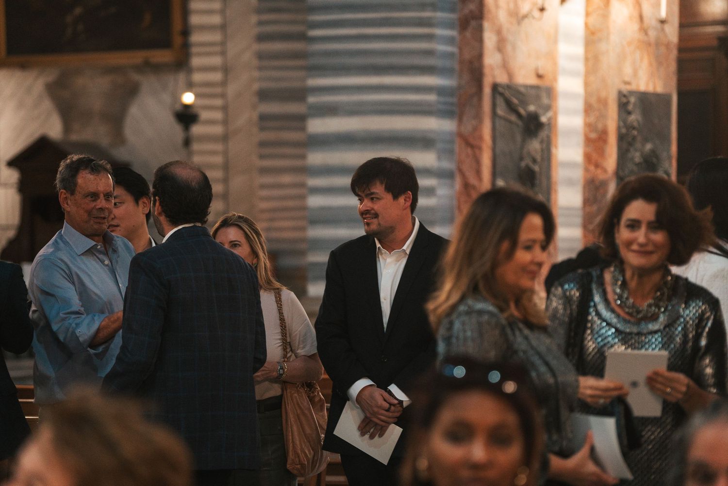 A crowded social gathering shows people mingling and conversing in an indoor venue with warm lighting.