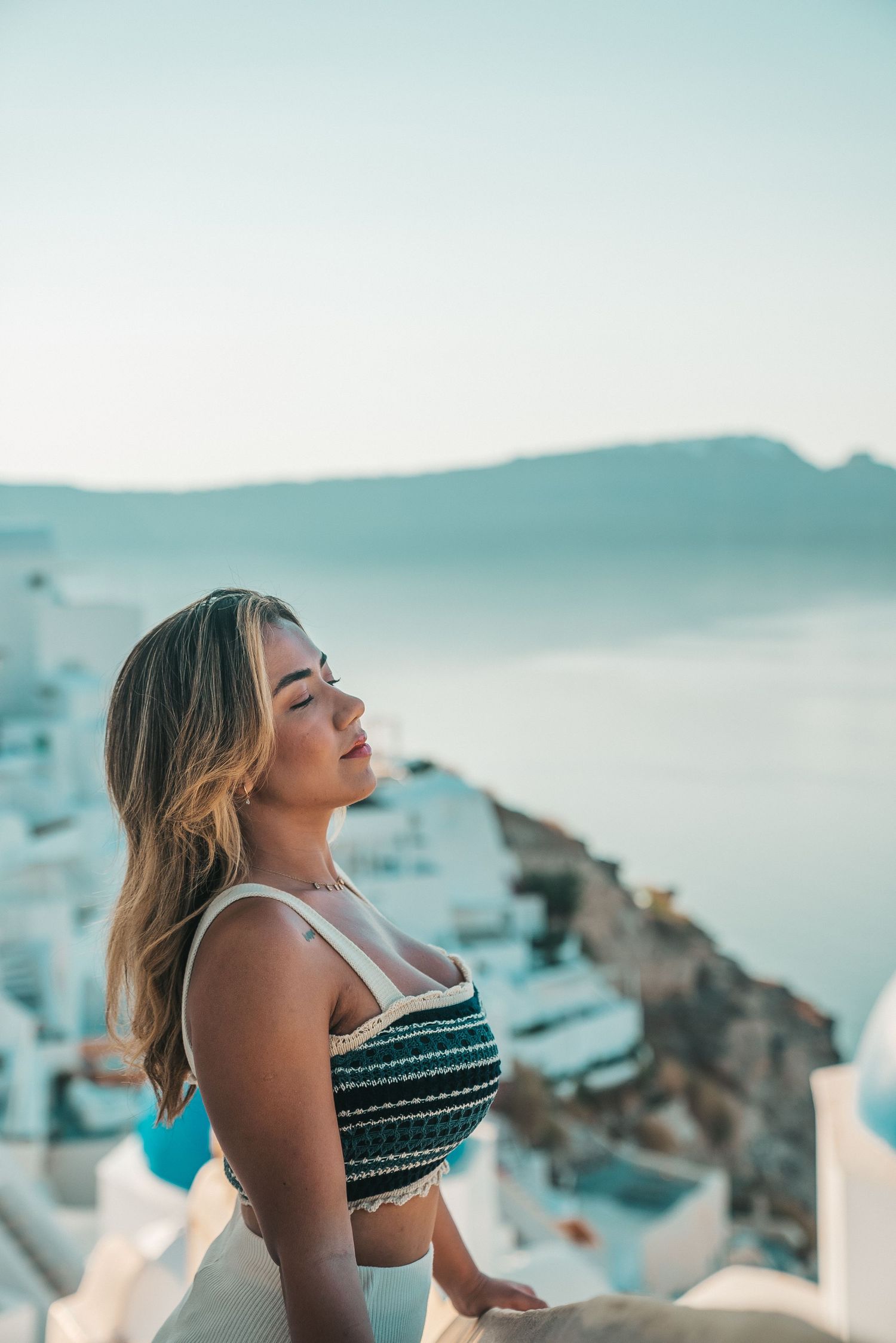 A person in a striped top enjoys the scenic coastal view of Santorini, Greece at sunset with white buildings visible.