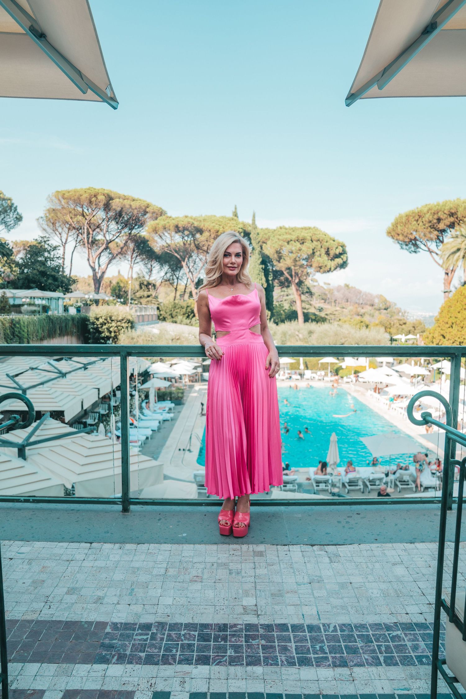A pink evening gown on a balcony overlooking a resort swimming pool with Mediterranean pine trees in the background.