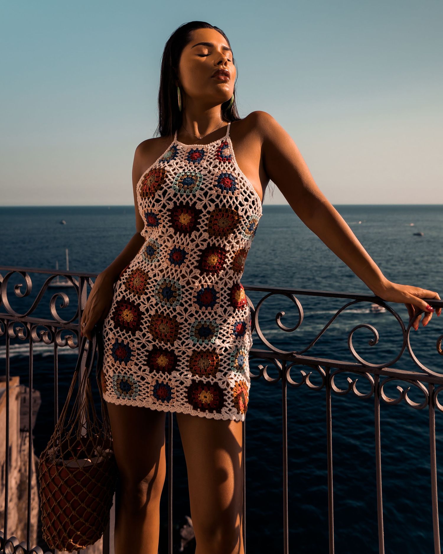 A person in a crochet dress poses against an ornate railing overlooking the ocean at sunset.