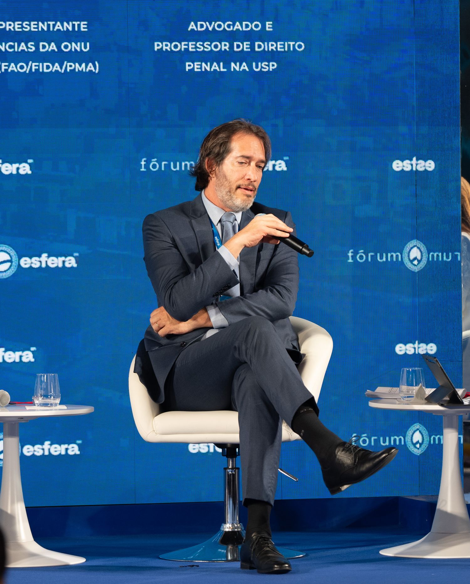 Speaker sits in white chair against blue backdrop during panel discussion at professional forum event.