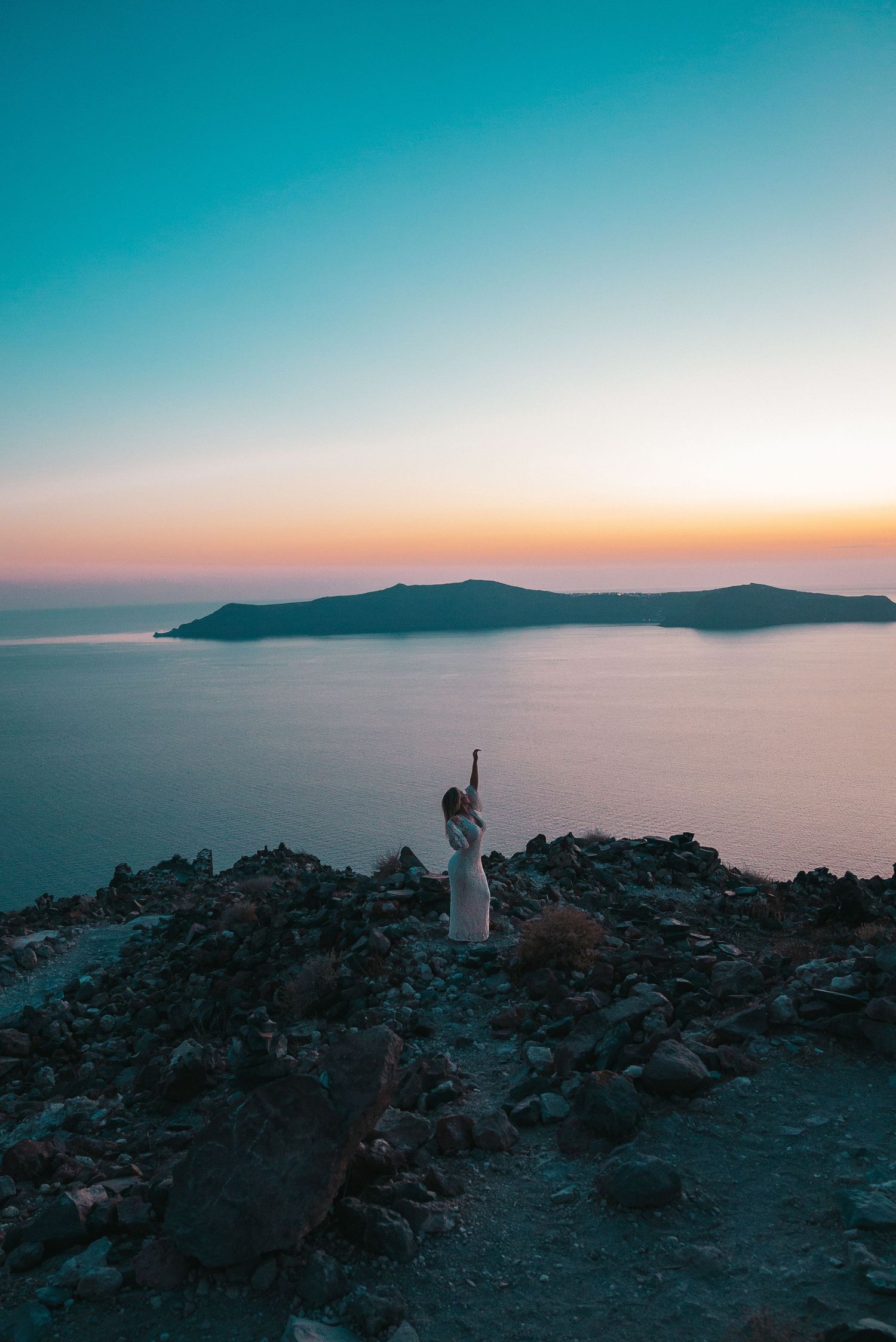 Silhouetted figures stand on rocky cliff overlooking a serene sunset ocean view with mountains in the distance.