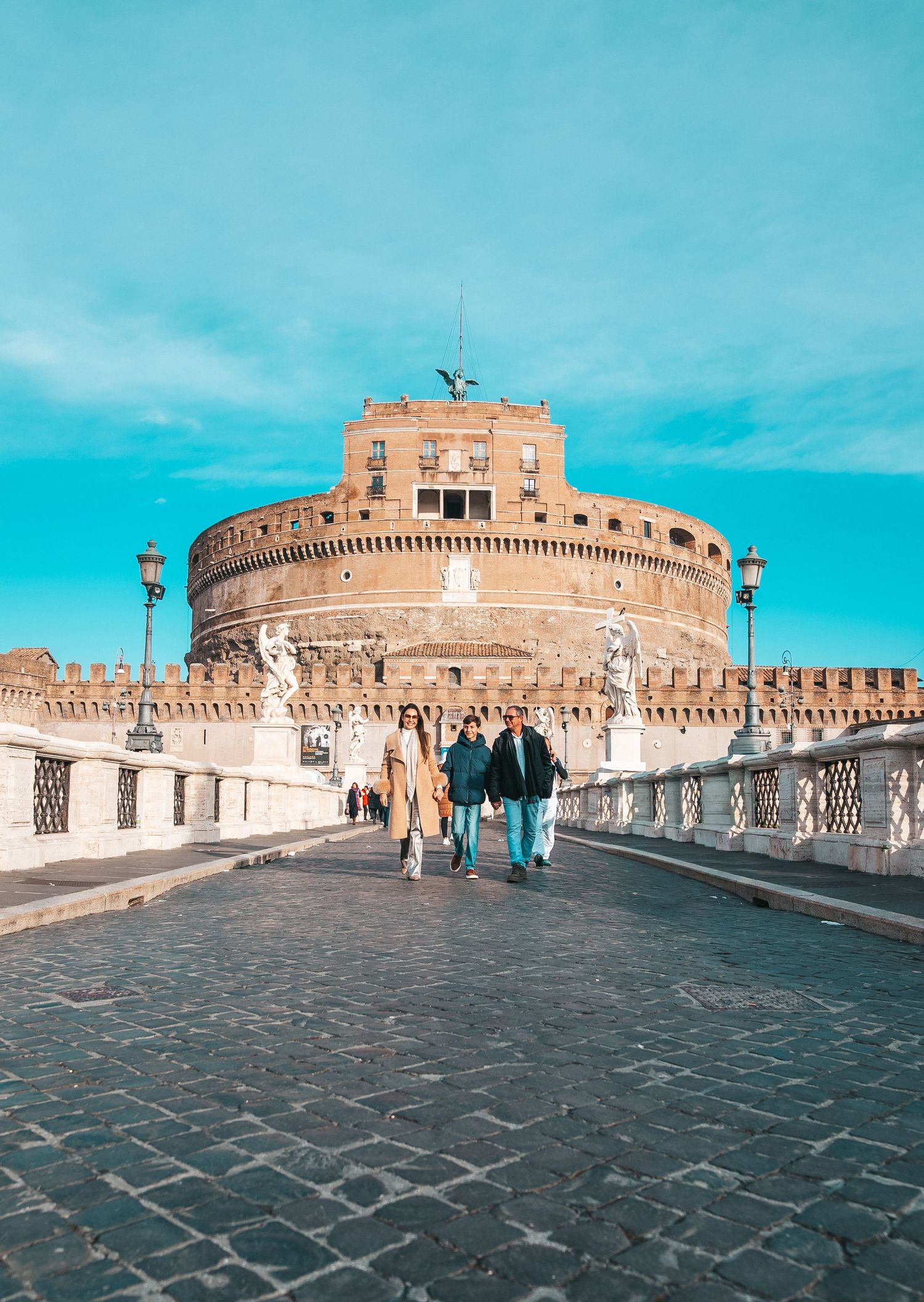 Castel Sant'Angelo in Rome with its iconic circular fortress and angel statues along the bridge under blue skies.