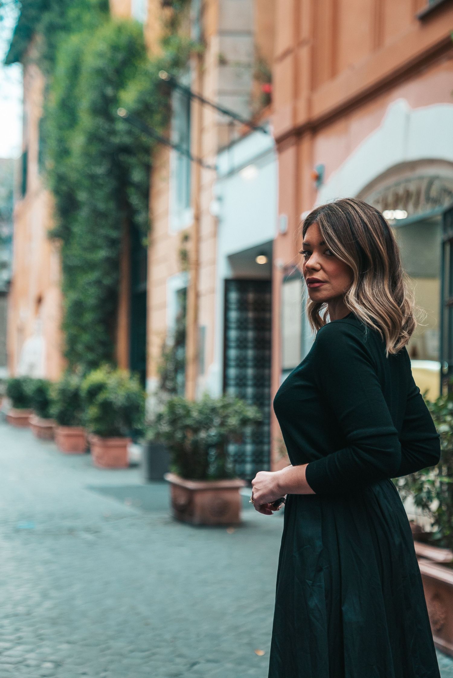 Person in black dress walking along cobblestone street with ivy-covered buildings.