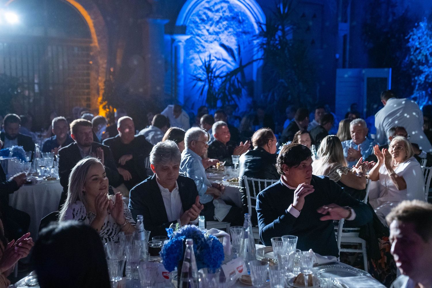 Elegant evening event with guests seated at tables in a dimly lit venue with blue architectural lighting.