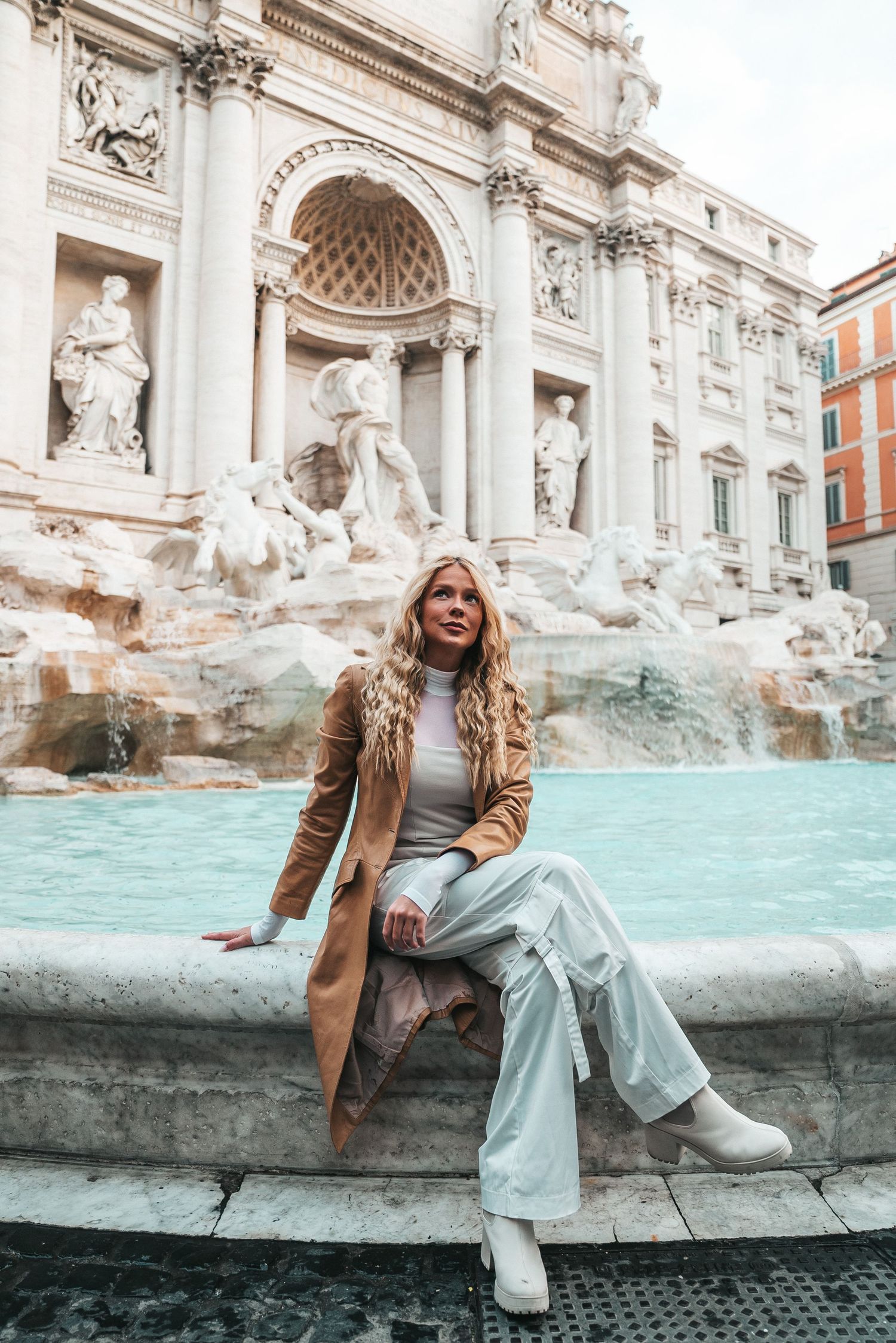 Tourist sitting on edge of Trevi Fountain in Rome wearing tan coat and light colored pants.