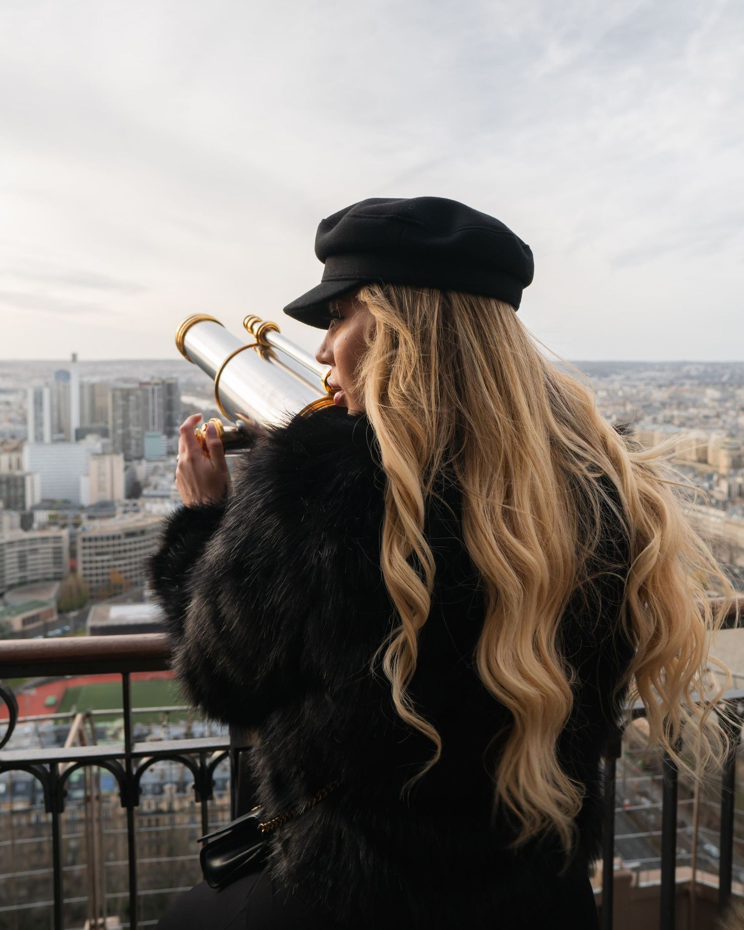 A person with blonde hair wearing a black hat looks out at a city skyline while drinking coffee on a balcony.