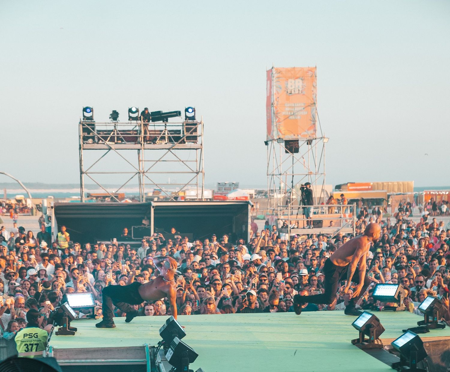 A large crowd watches performers on an outdoor stage at a music festival with scaffolding and equipment visible.