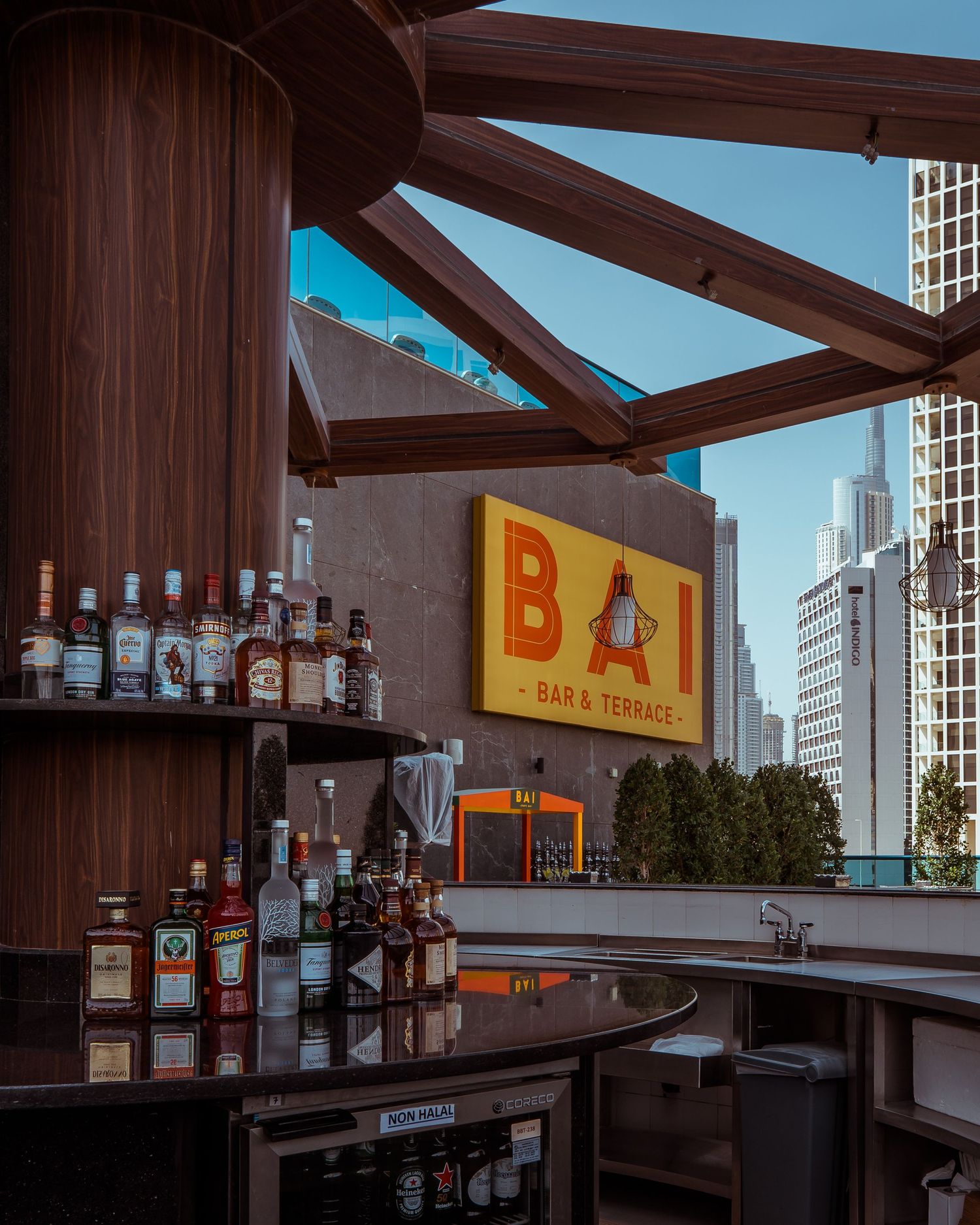 Rooftop bar with wooden beams and liquor bottles displayed against a blue sky with city buildings in background.