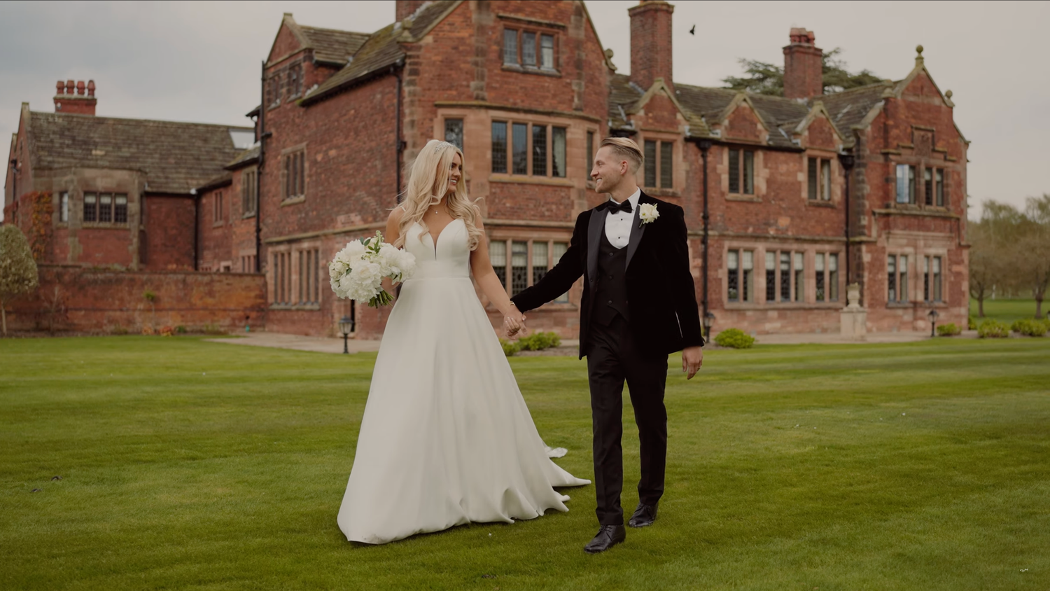 Elegant wedding couple walks across a lush green lawn in front of a historic red brick English manor house.