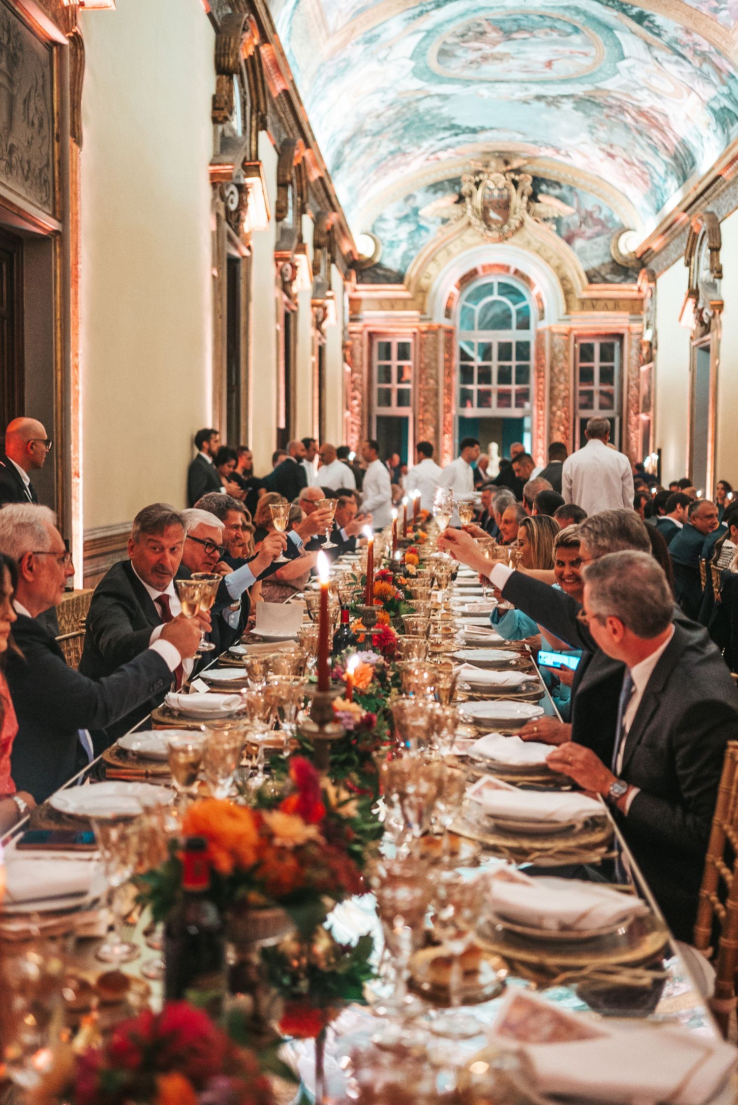 Elegant formal dinner setup in ornate hall with vaulted painted ceilings and long tables set with fine china and floral decor.