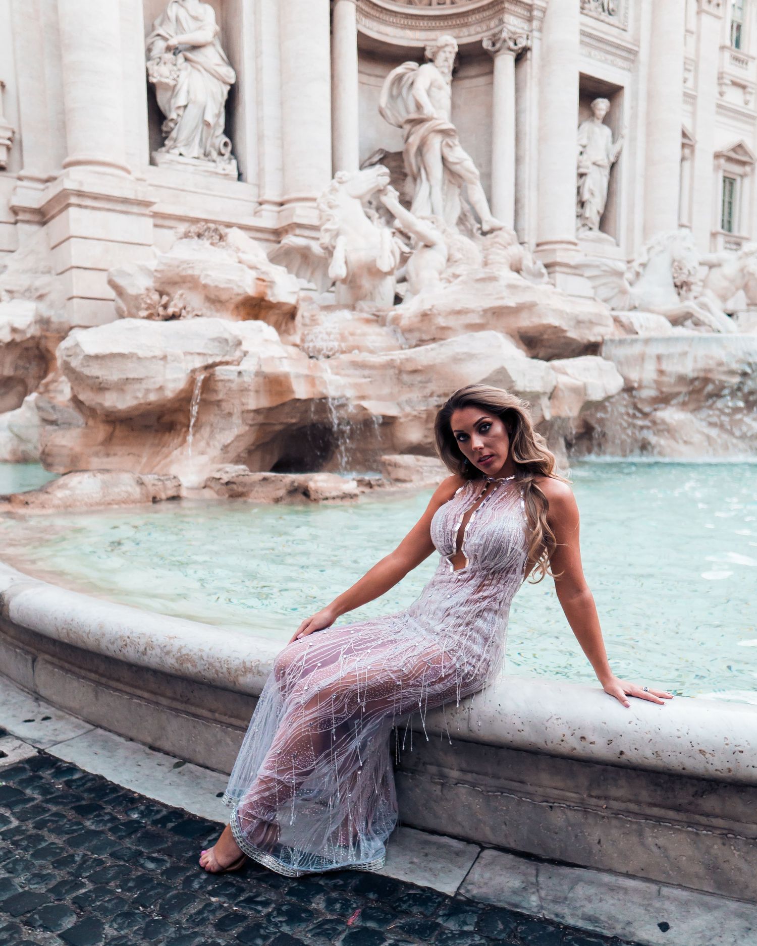 A person in a flowing lace dress poses gracefully by the edge of the Trevi Fountain in Rome, Italy.