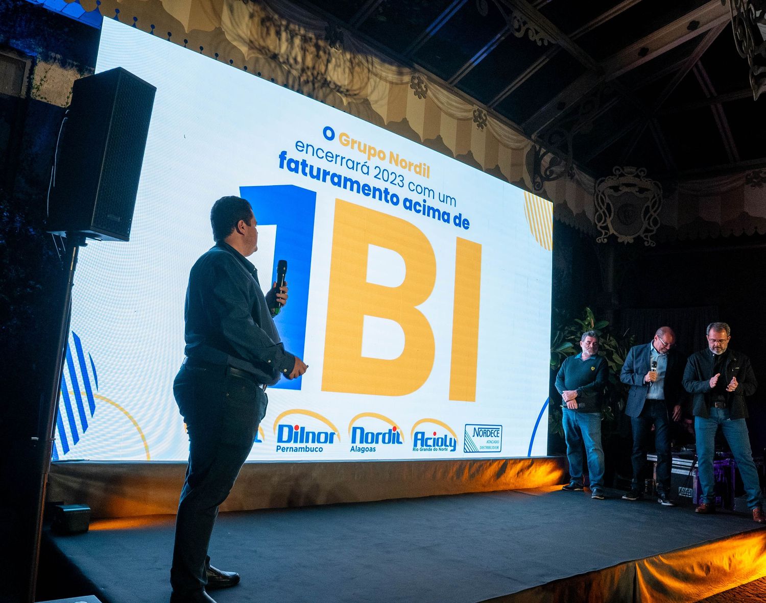 Speaker presenting on stage at a business event with large yellow and blue IBI logo projected on wall.