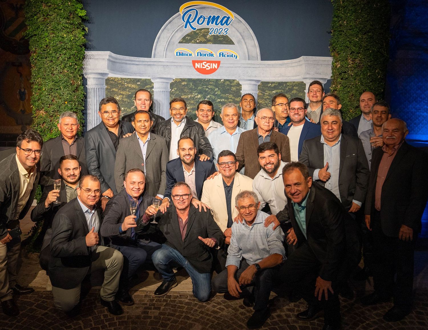 Large group of people posing together for a team photo in front of a Roma logo backdrop.