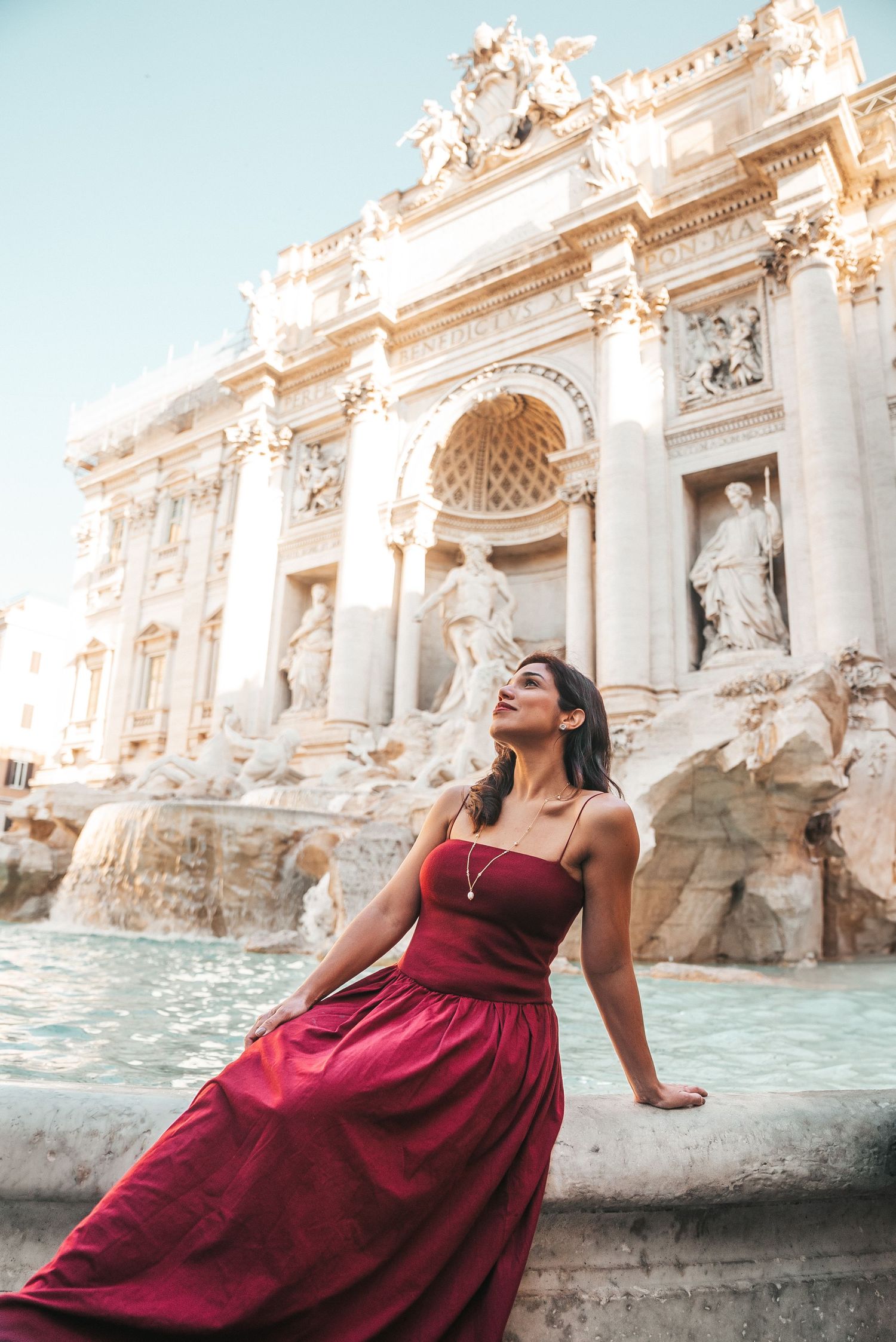 A woman in a burgundy dress poses elegantly by the ornate Trevi Fountain in Rome, Italy.