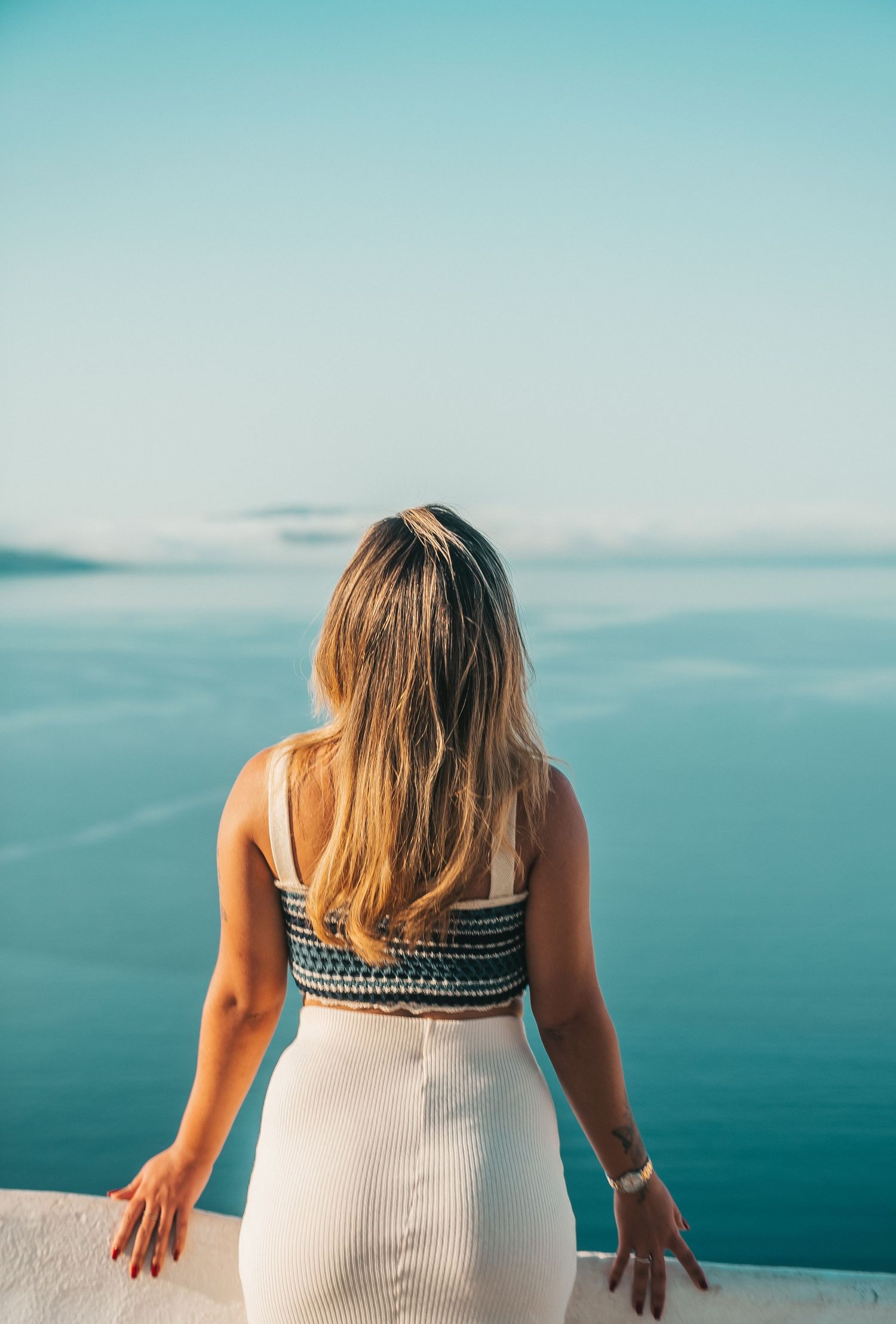 Person in white dress sits overlooking a serene blue ocean view against clear sky on sunny day.