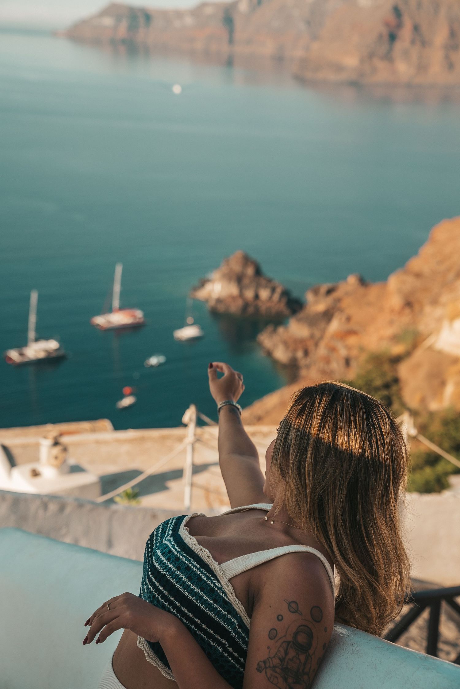 Person admiring scenic Santorini coastline with sailboats and cliffs from a balcony viewpoint at sunset.