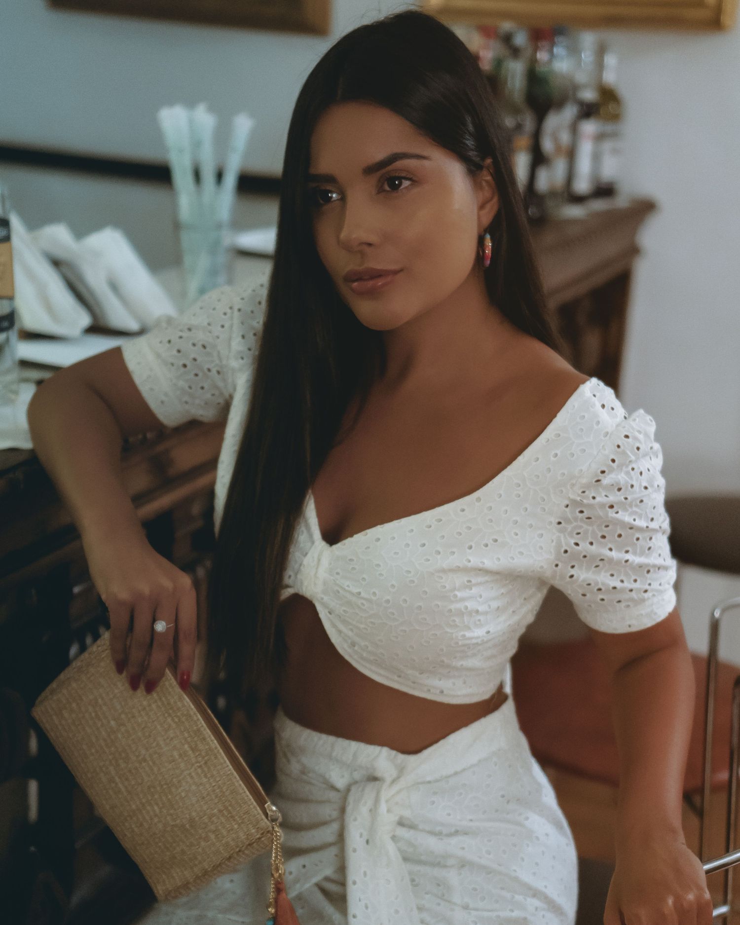 Woman in white crop top with puff sleeves and high waisted pants holds a woven clutch purse in indoor setting.