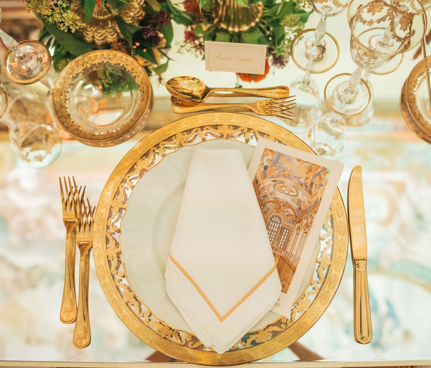 Elegant gold-rimmed place settings with crystal stemware and ornate details on a white tablecloth for formal dining.