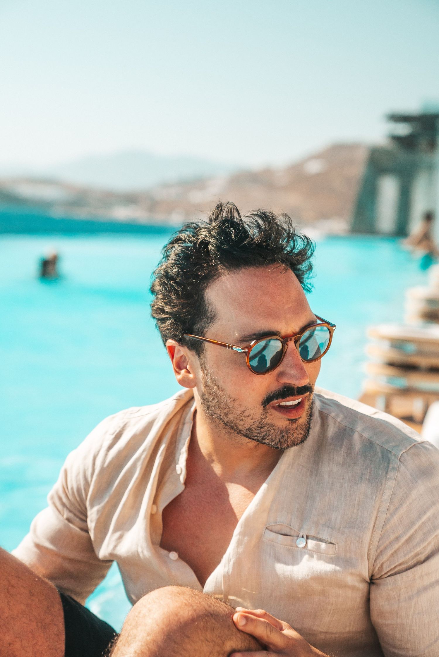 Someone wearing sunglasses and a linen shirt relaxes by a bright blue pool with mountains in the background.