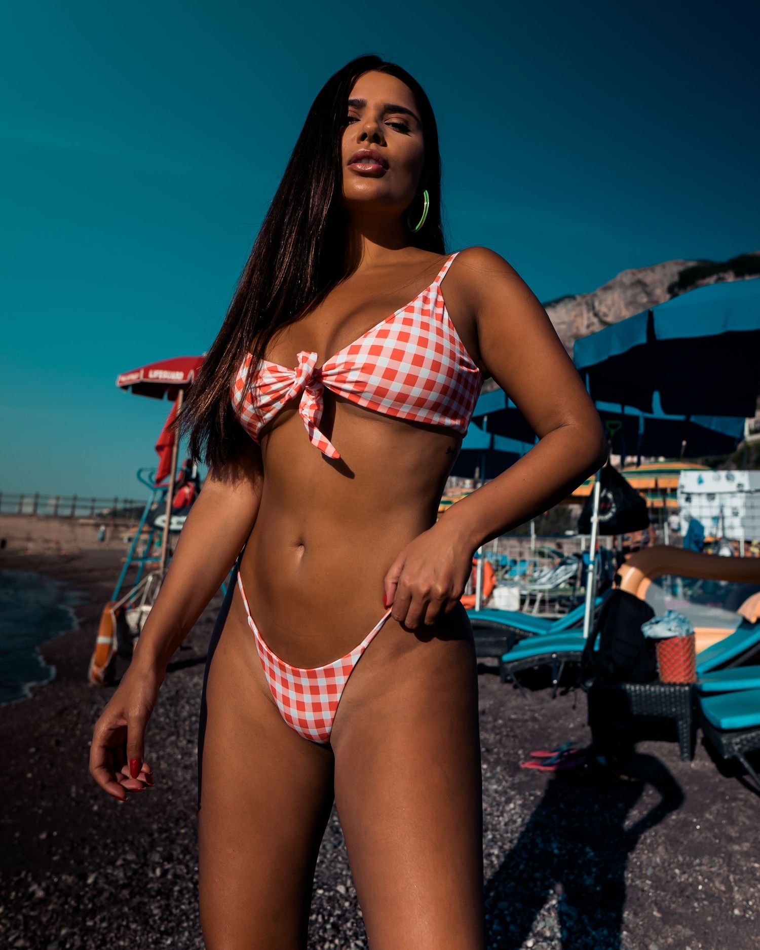 Person in red and white gingham bikini poses on dark rocky beach against vibrant blue sky background.